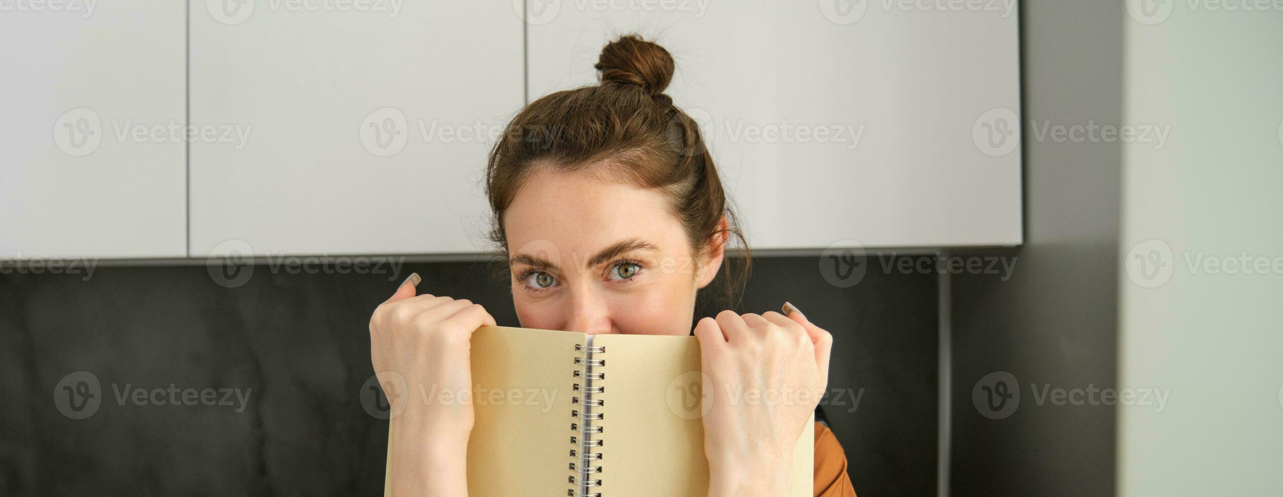 estilo de vida y personas concepto. joven morena mujer, participación cuaderno cerca rostro, ocultación sonrisa detrás bloc y mirando jugado a cámara foto