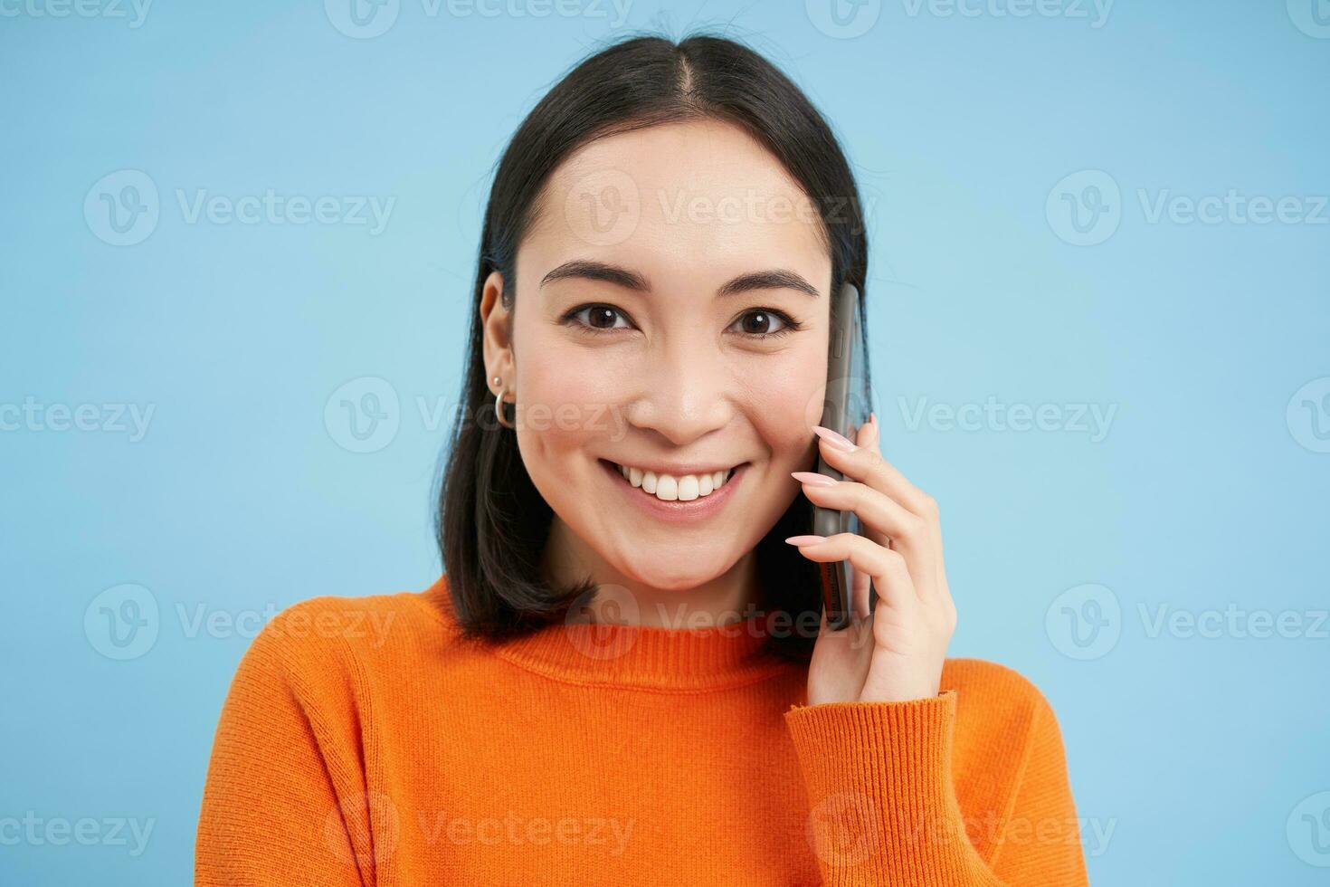 Technology concept. Close up of smiling asian woman talks on mobile phone, having conversation on cellphone, blue background photo