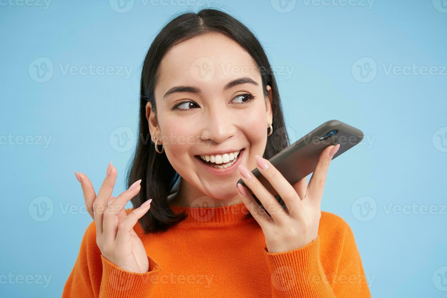 Portrait of asian woman records voice message, talks on speakerphone, holds mobile phone near mouth while speaking, translates her speech on app, blue background photo