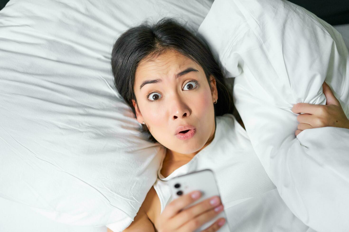 Portrait of asian girl wakes up in morning, looks at mobile phone with shocked face, overslept, hugging pillow and grimacing photo
