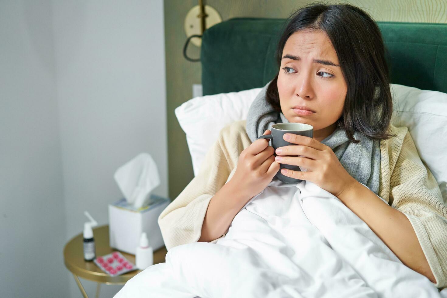 Covid-19, flu and vaccination concept. Korean girl lying in bed with cold, catching influenza, drinking hot tea and using prescribed medication photo