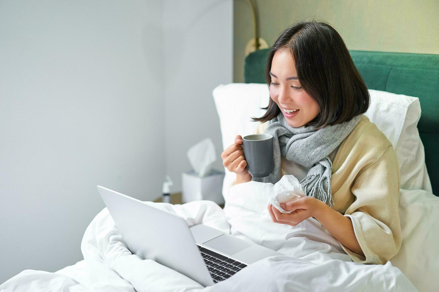 Portrait of asian sick girl, lying in bed with cold, having flu, talking to laptop, video chat, working from home on remote while on sick leave photo
