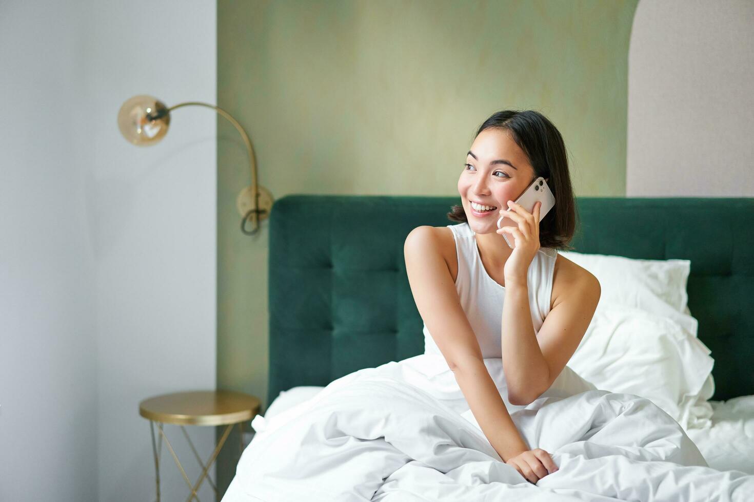 Beautiful smiling asian girl talking on mobile phone, lying in bed with pleased happy face, speaking to someone on telephone photo