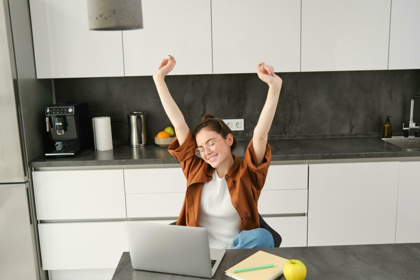 Productivity and lifestyle concept. Beautiful young woman finishes working, stretching hands with satisfied expression, sitting in kitchen with laptop and documents, has remote workplace at home photo
