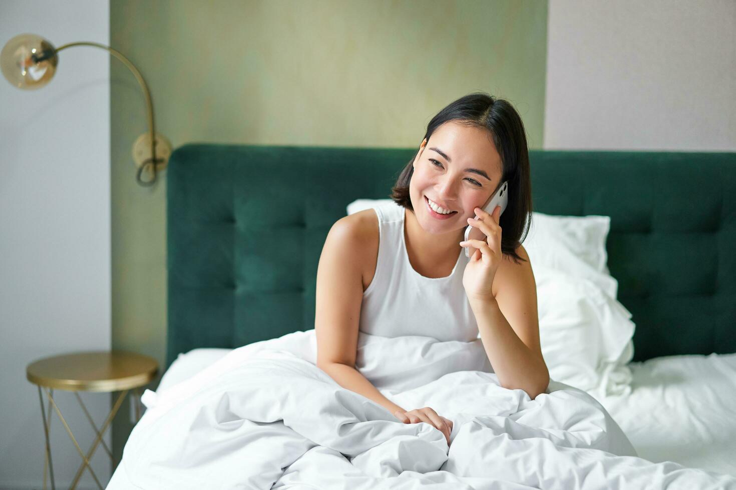 hermosa joven coreano mujer vocación, teniendo un teléfono llamada en cama, hablando con amigo mientras quedarse en dormitorio foto