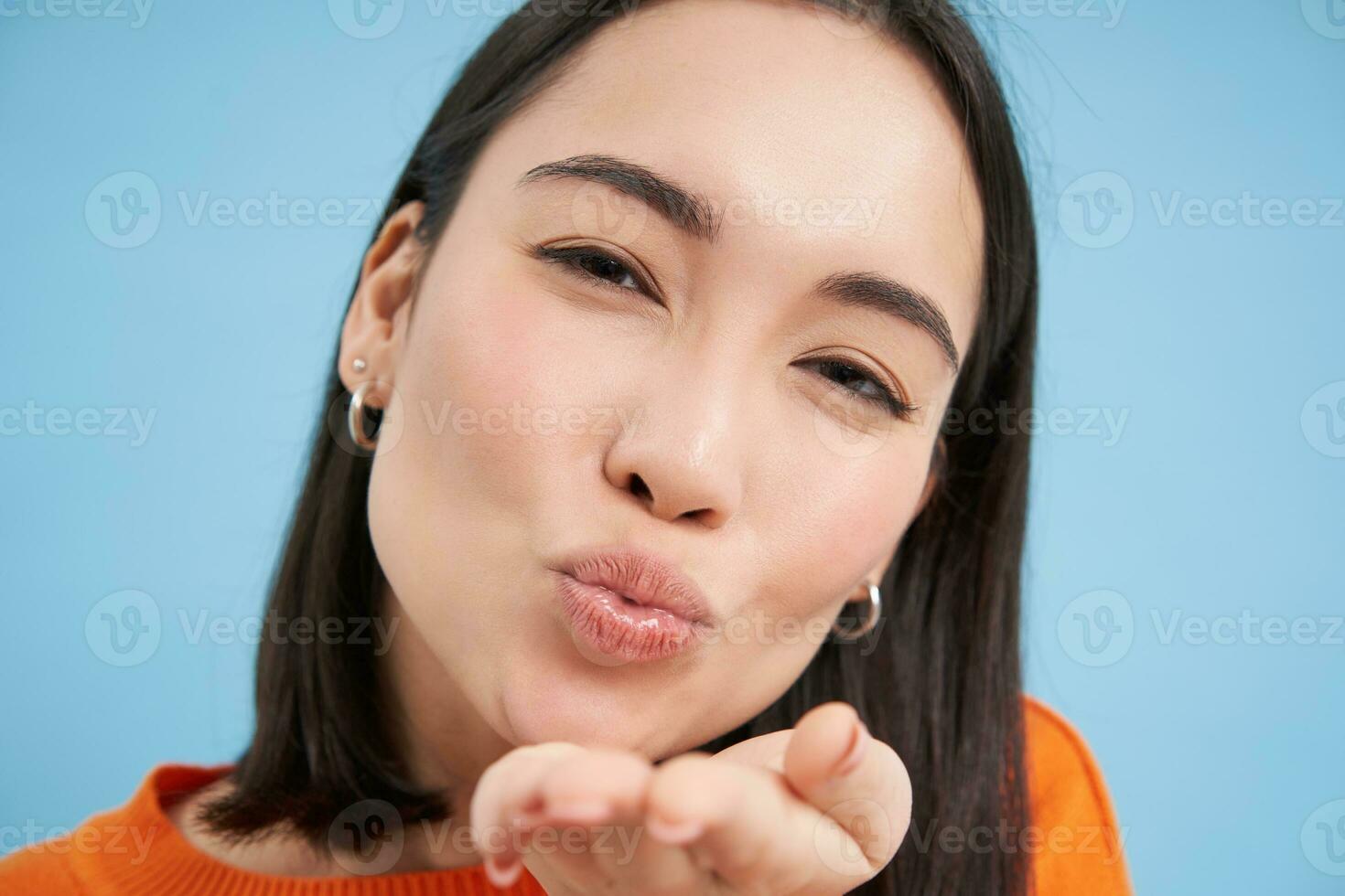 Close up of beautiful korean woman, blowing air kiss at camera, standing over blue background photo