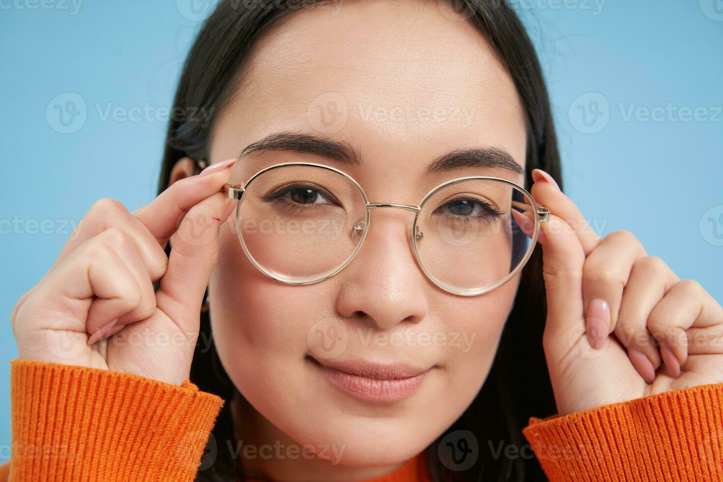 Close up portrait of amazed girl, looks closer at camera in glasses, standing against blue background photo