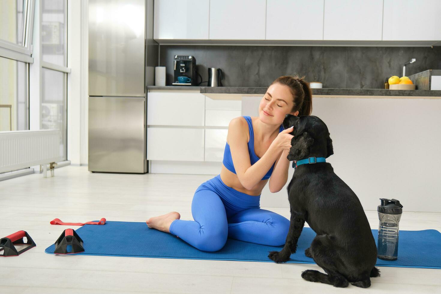 Portrait of beautiful brunette fitness girl, plays with her dog during workout training session, sits at home on rubber yoga mat, does sport exercises in living room photo