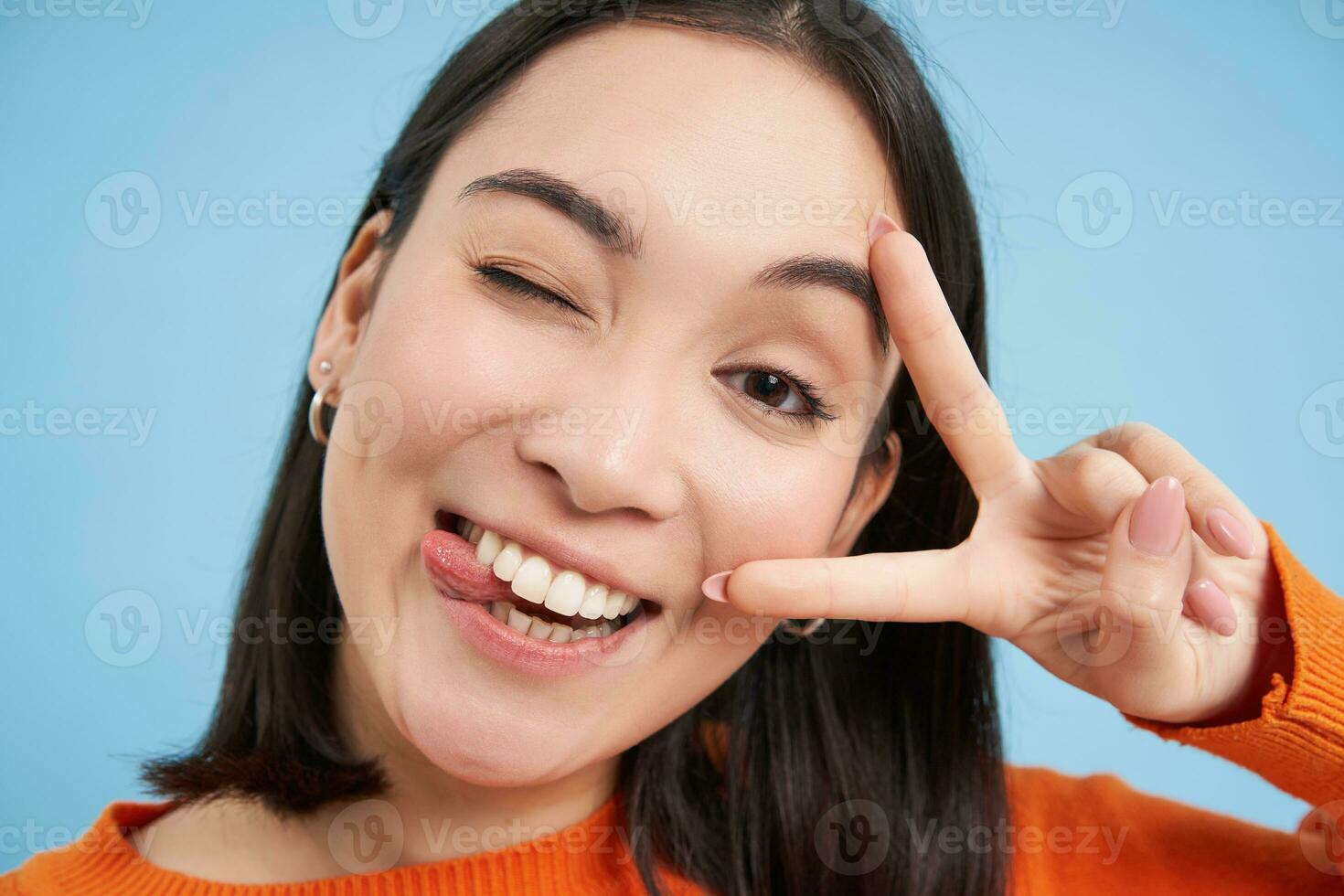 Close up portrait of positive asian woman, winks and shows peave, v-sign gesture, sticks tongue and smiles, stands over blue background photo