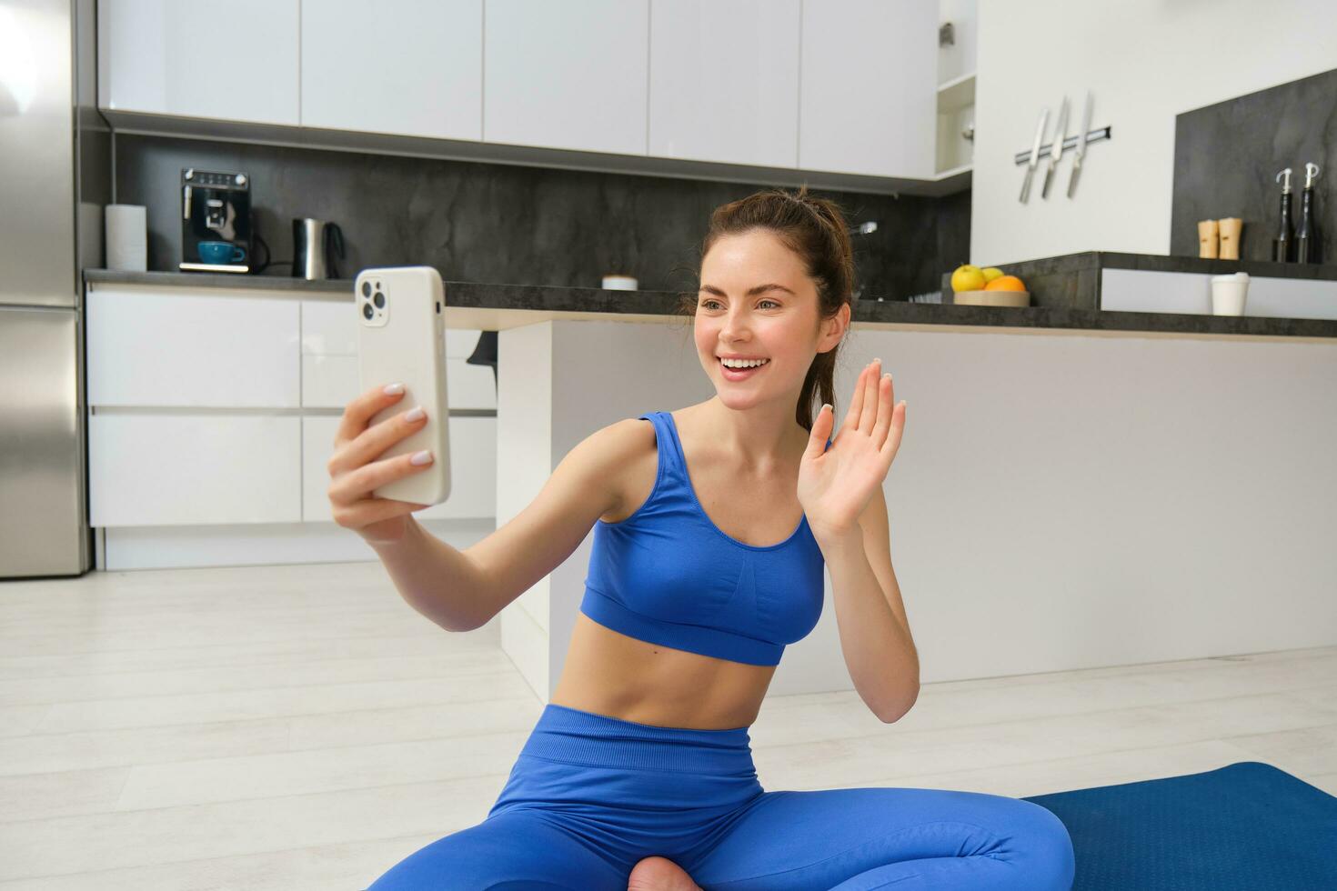 Portrait of young fitness girl, blogger records her workout from home, poses for selfie on smartphone, does yoga on camera, sits in blue sportsbra and leggings photo