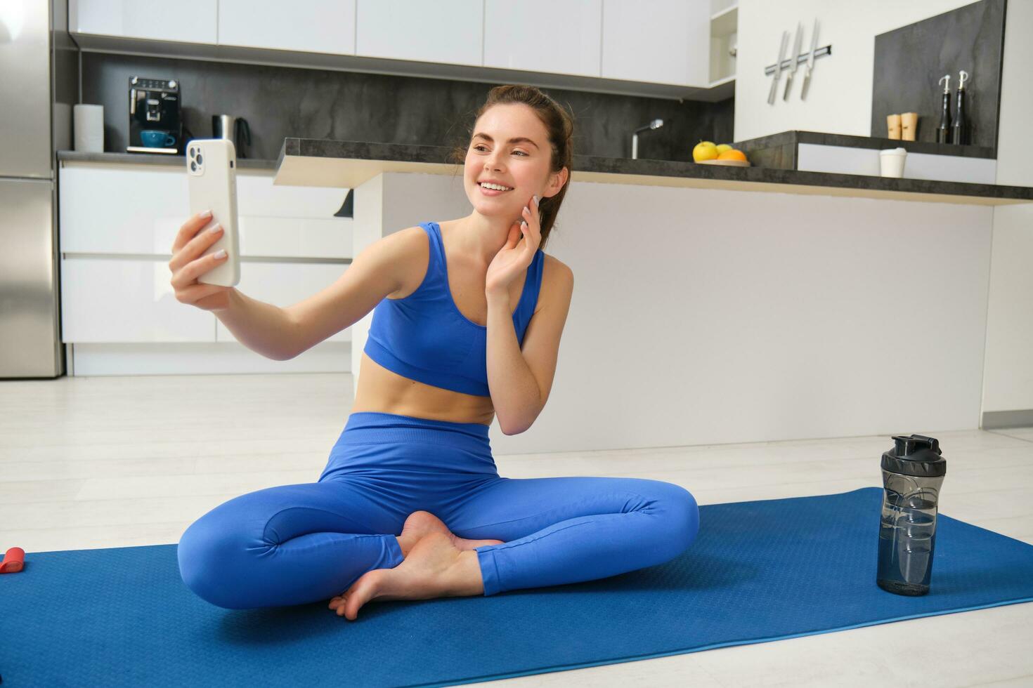 Portrait of woman workout from home, sitting on yoga mat with smartphone, taking selfie on mobile phone, doing exercises in her living room photo