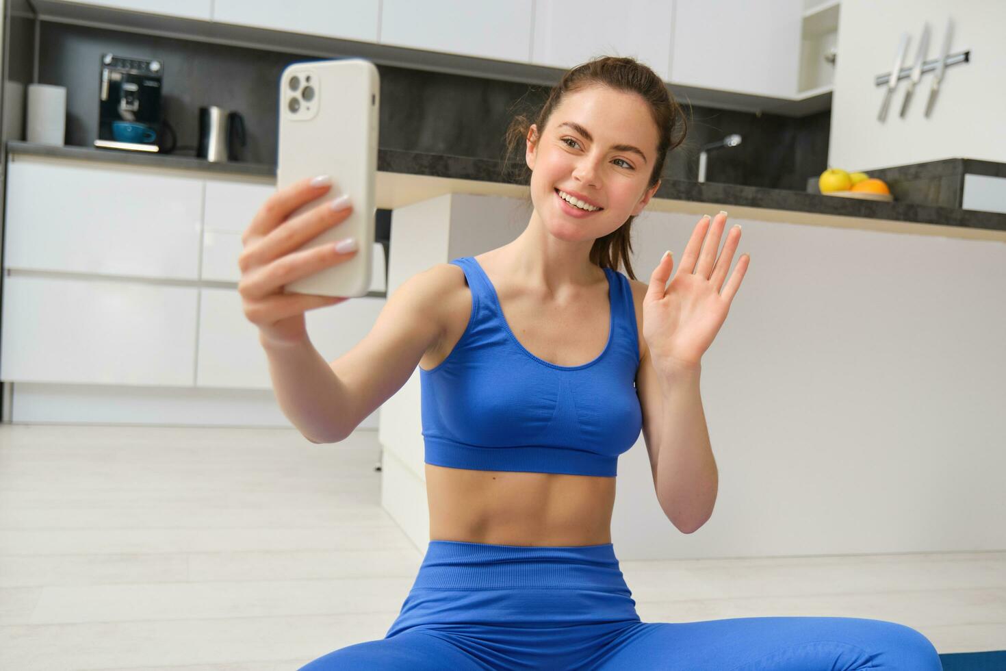 sonriente hermosa niña registros vídeo mientras haciendo Deportes a hogar, mira a teléfono inteligente, toma selfie en móvil teléfono, rutina de ejercicio adentro en azul polainas y sujetador deportivo foto