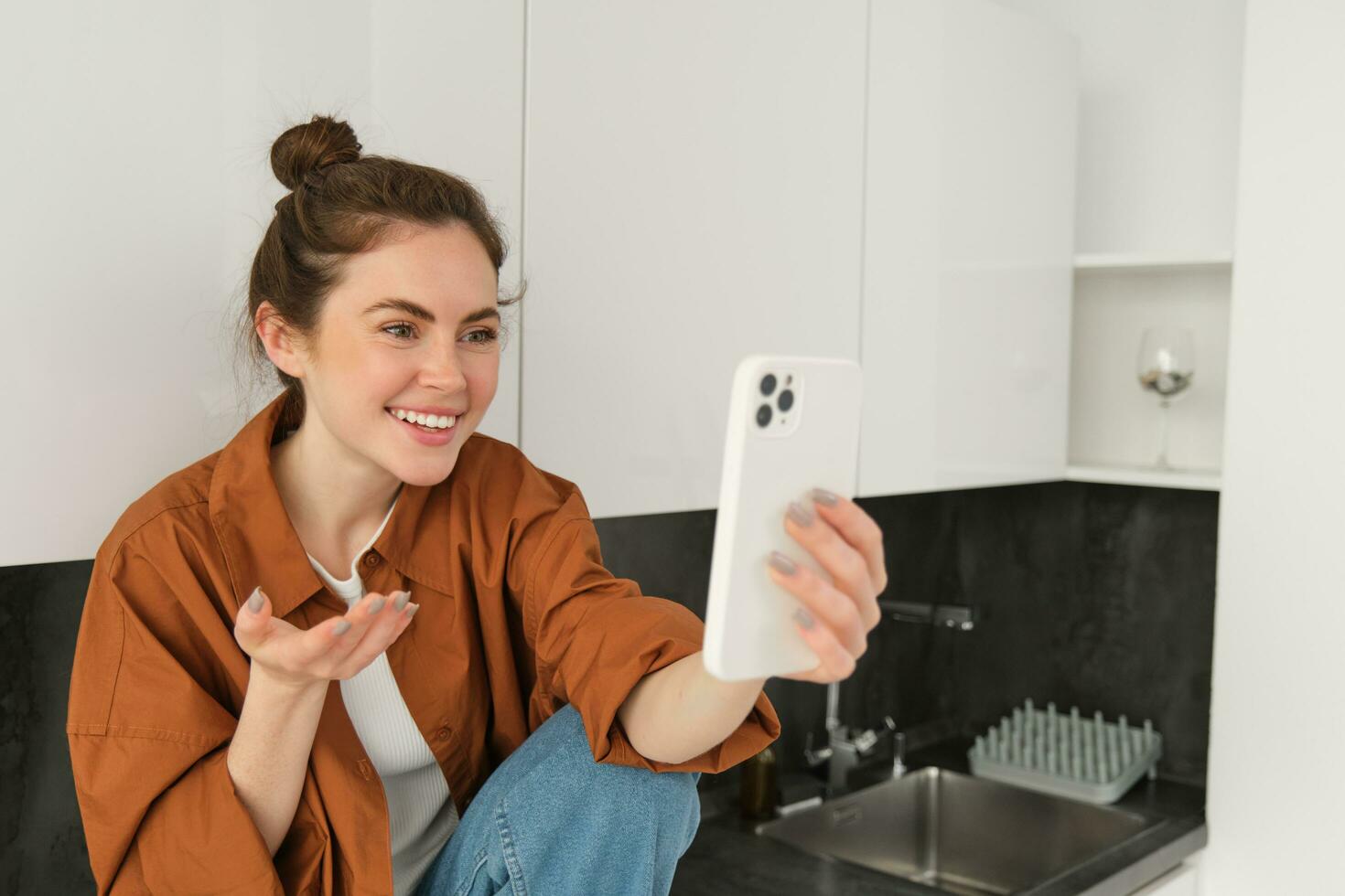 Portrait of young cheerful woman laughing and smiling during phone call, video chats with friend, sits on kitchen counter and talks to someone using smartphone app photo