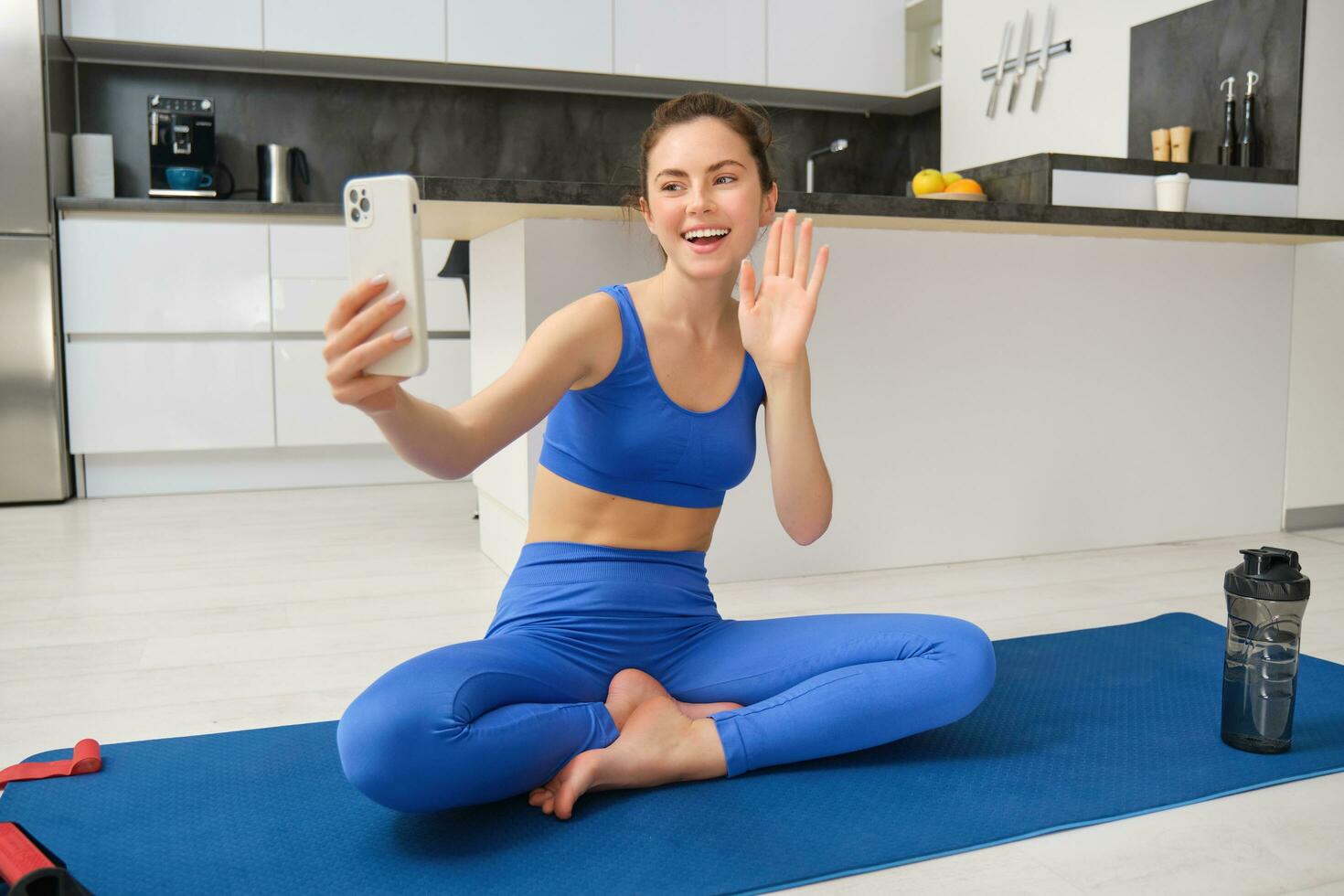 vlogger lo hace Deportes, registros su rutina de ejercicio formación desde hogar en teléfono inteligente cámara, posando para selfie dentro su casa, se sienta en caucho yoga estera en azul polainas y sujetador deportivo foto