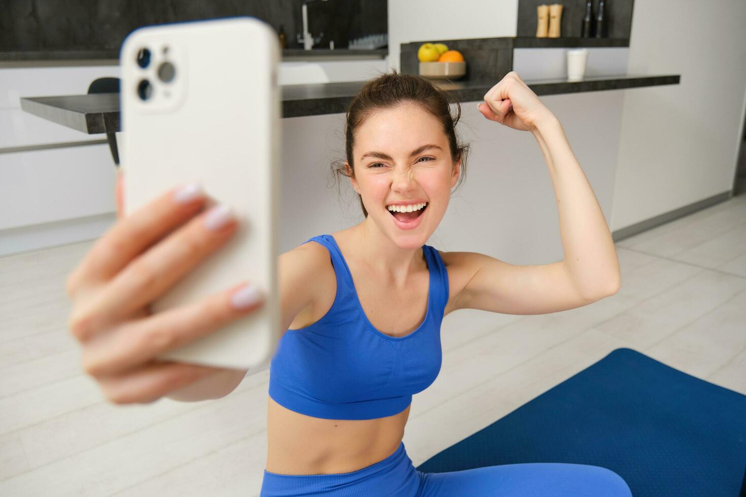 Young energetic girl does sports at home, shows her muscles on arm and takes selfie on smartphone, posing on fitness rubber mat in living room photo