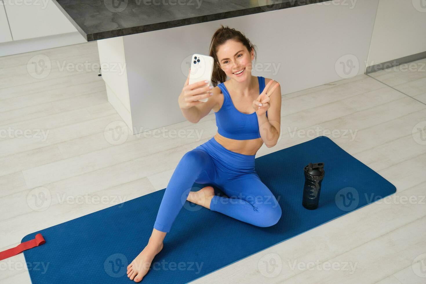 Portrait of stylish, smiling young brunette woman, wears blue sportswear, poses on yoga mat with smartphone, shows peace sign and takes selfie, does workout at home photo