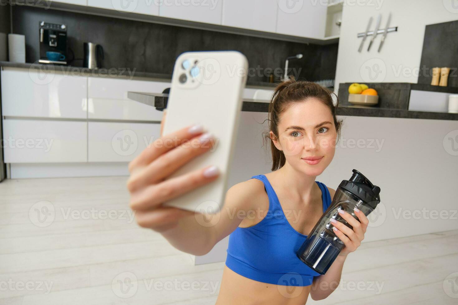 Portrait of young fitness girl, takes selfie on smartphone with water bottle, drinks water during workout, training at home, records sports vlog photo