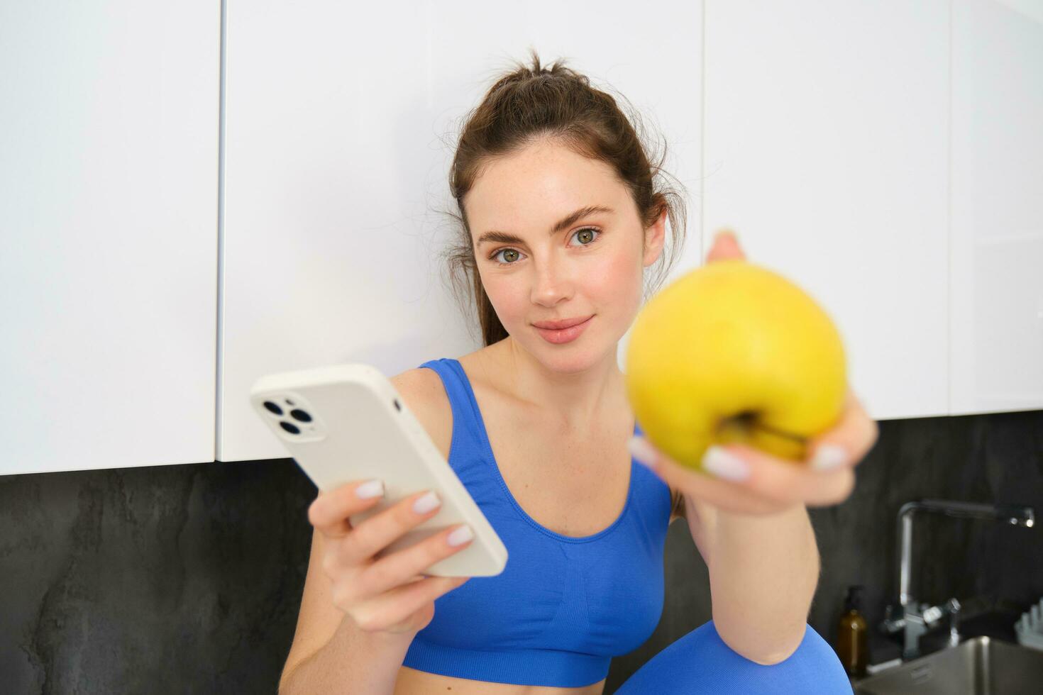 Image of beautiful, sportswoman sitting in kitchen with smartphone, offering you an apple, eating fruits, wearing sportsbra and leggings photo