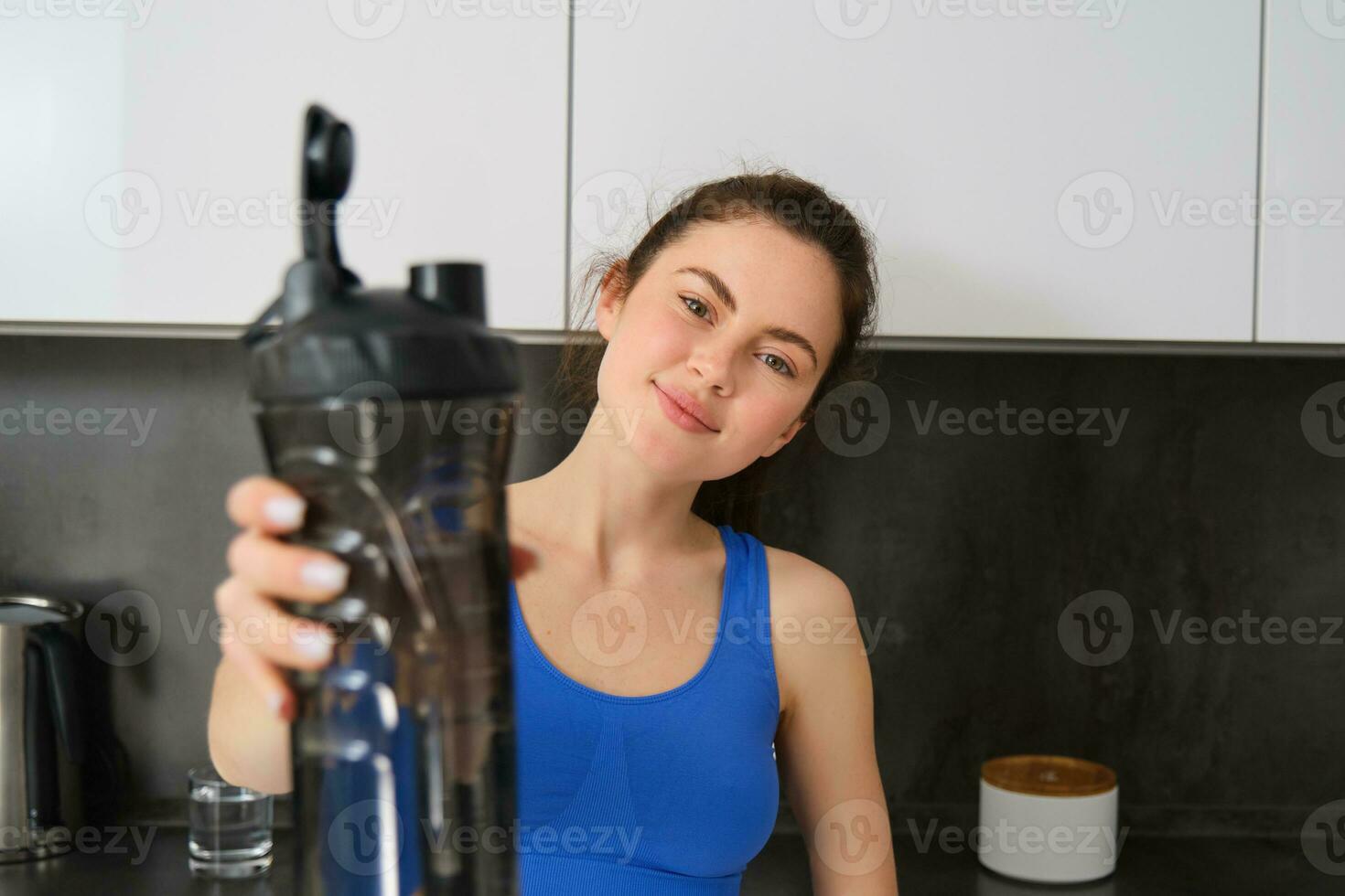 retrato de hermosa joven aptitud chica, ofrecimiento usted agua botella a bebida después ejercicio, en pie en cocina, sonriente a cámara foto