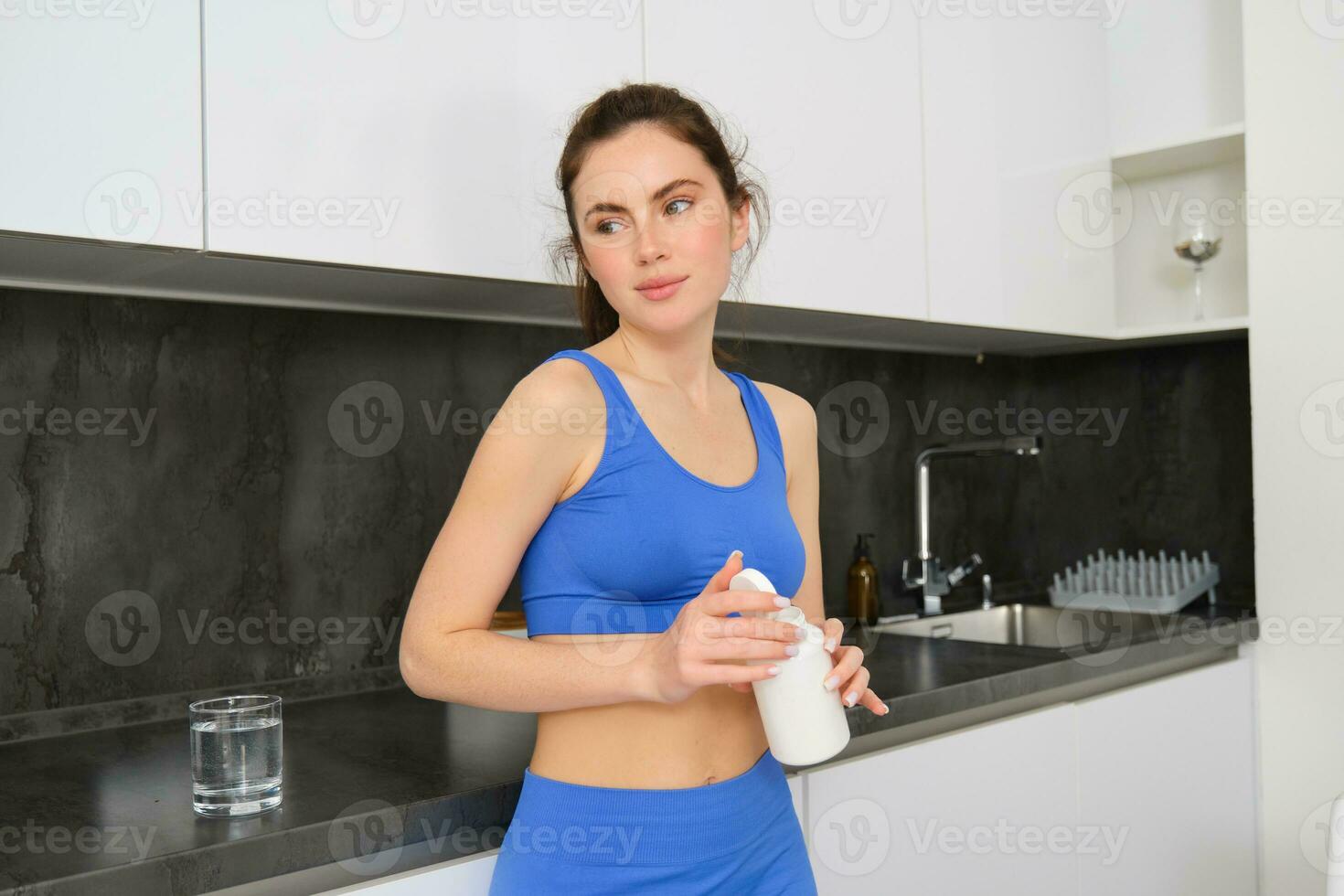 Portrait of healthy, beautiful woman, holding bottle of vitamins, taking dietary supplements, buds for shiny skin and strong muscles, standing in fitness clothing photo