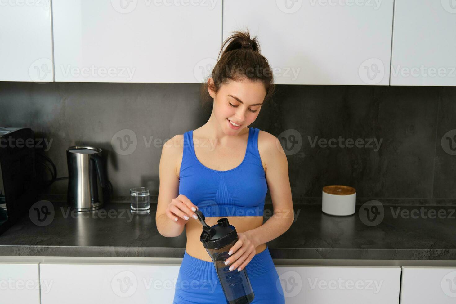 imagen de joven mujer preparando dietético suplemento, proteína en su criba vibradora botella, vistiendo aptitud ropa, en pie en cocina foto