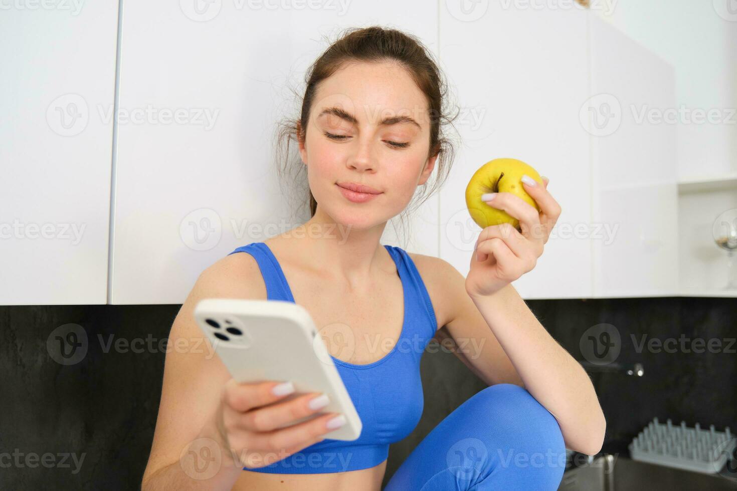 Portrait of sportswoman, girl eating and apple and looking at her social media, smartphone screen, having a snack in kitchen, wearing fitness activewear photo