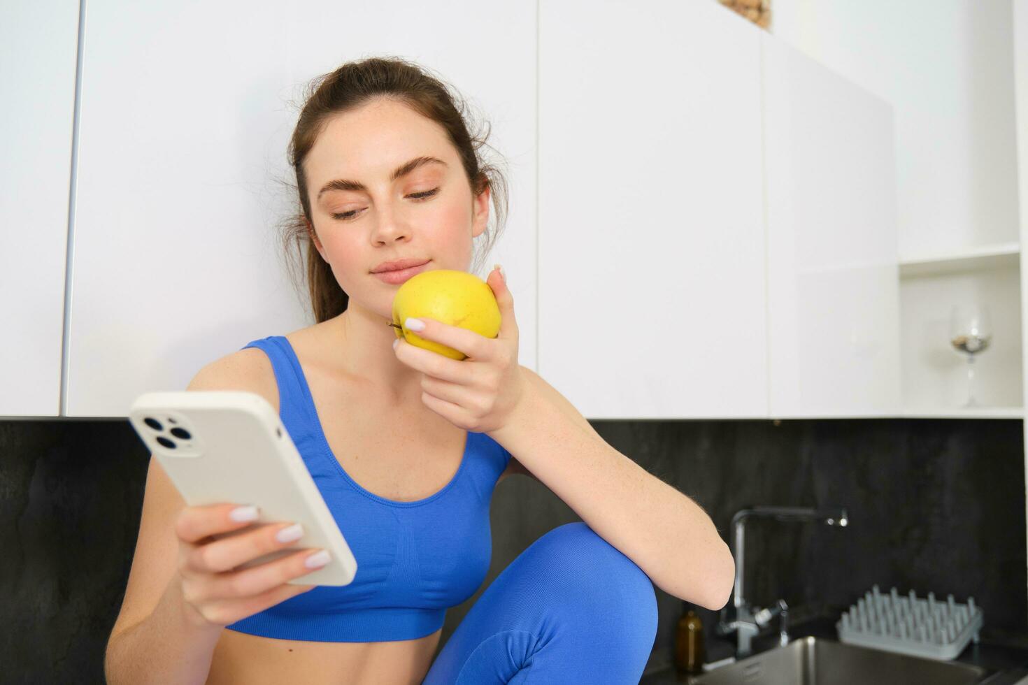 Image of stylish sportswoman, young fitness instructor sitting in kitchen and eating an apple, holding smartphone, using social media app on mobile phone photo