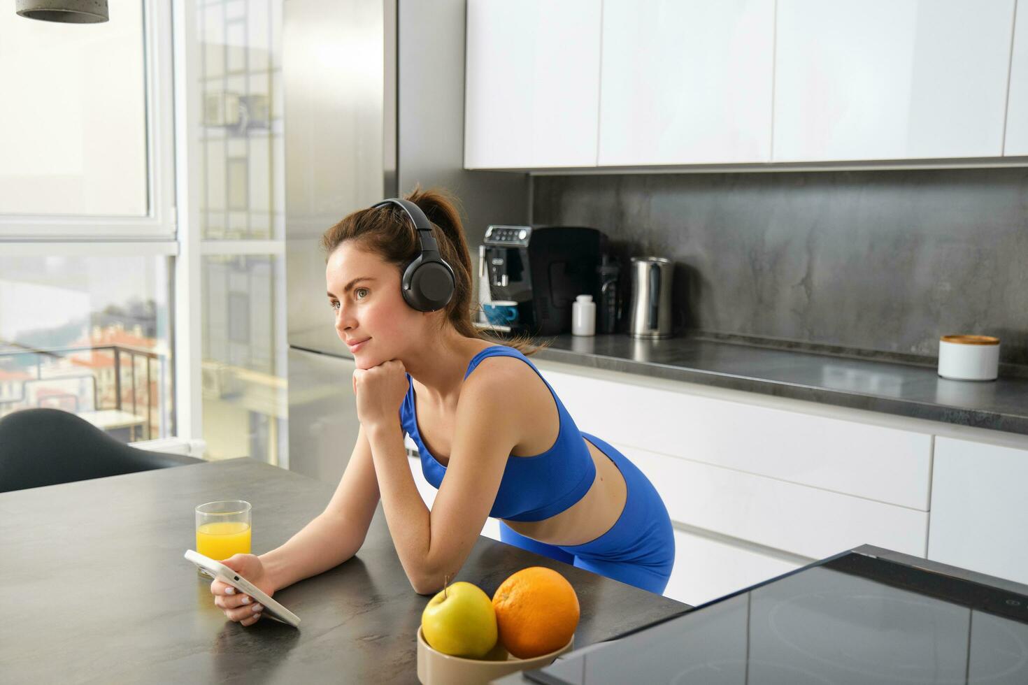 Portrait of stylish sportswoman, listening music in headphones, looking at smartphone, watching video on mobile phone, standing in kitchen, wearing activewear photo