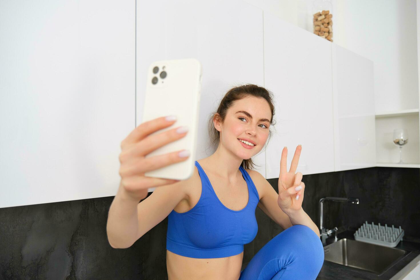 Portrait of beautiful woman, fitness blogger, taking selfie in sportsbra and leggings, posing in kitchen with smartphone photo