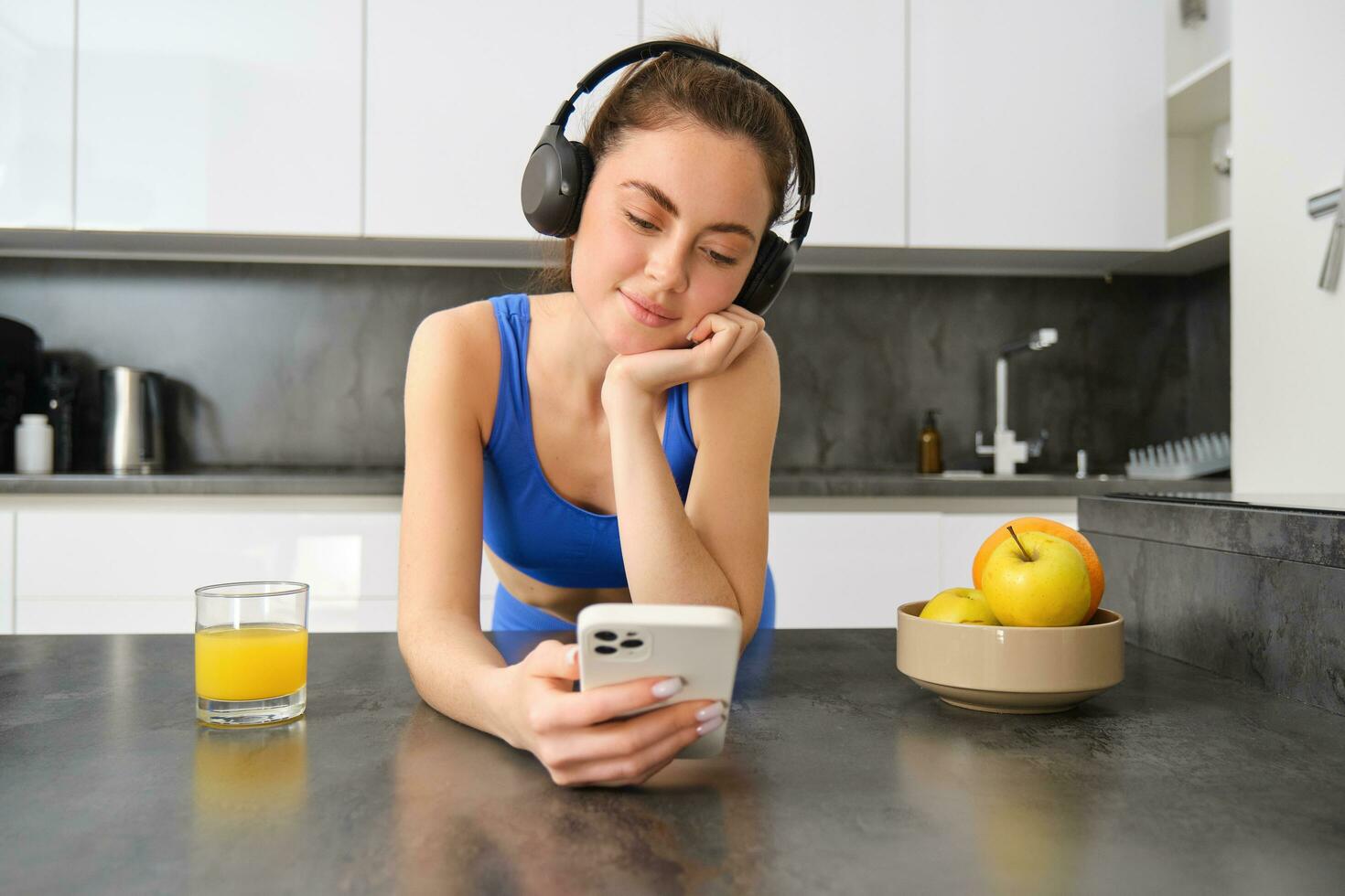 Lifestyle and workout. Young smiling woman in headphones, standing in kitchen with smartphone, drinking orange juice and listening music, heading over to gym, going jogging photo