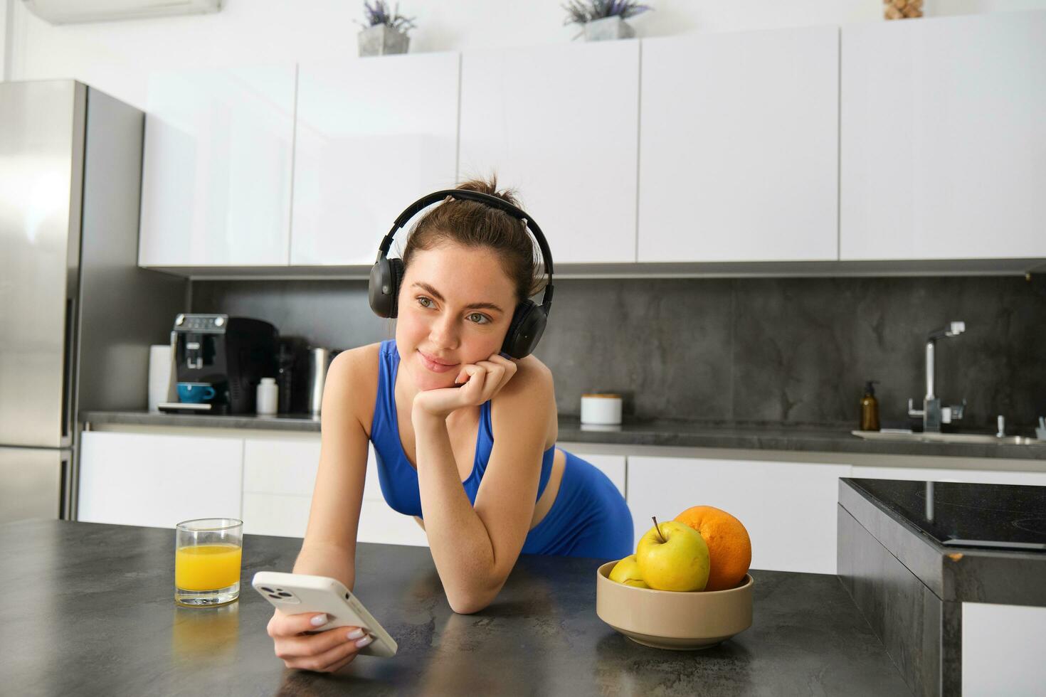 Portrait of young fitness woman with headphones, drinking orange juice in kitchen and using smartphone, listening music, getting ready for workout gym photo