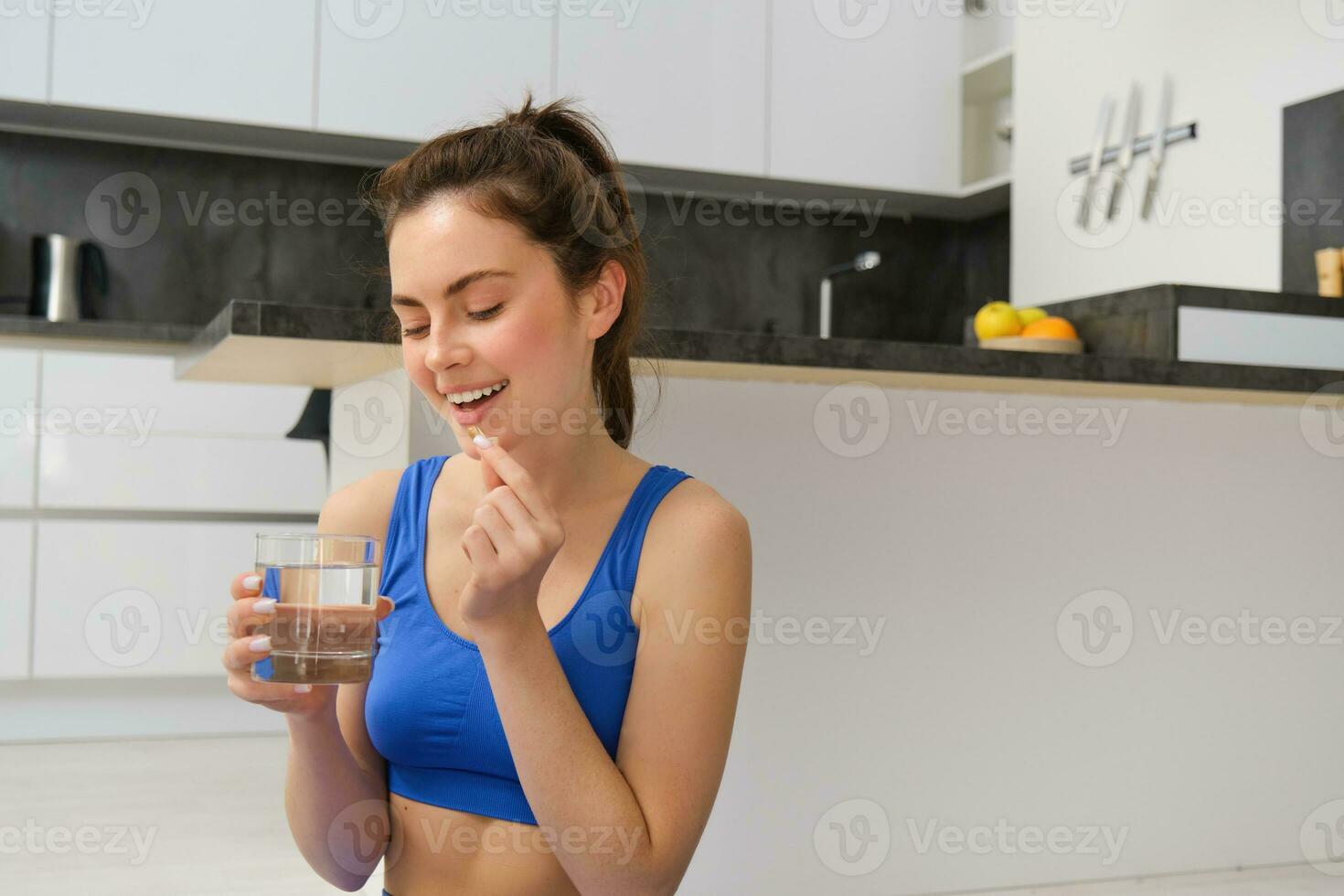Portrait of healthy, smiling young woman taking vitamins after workout, holding glass of water and dietary supplement, buds for health and energy photo