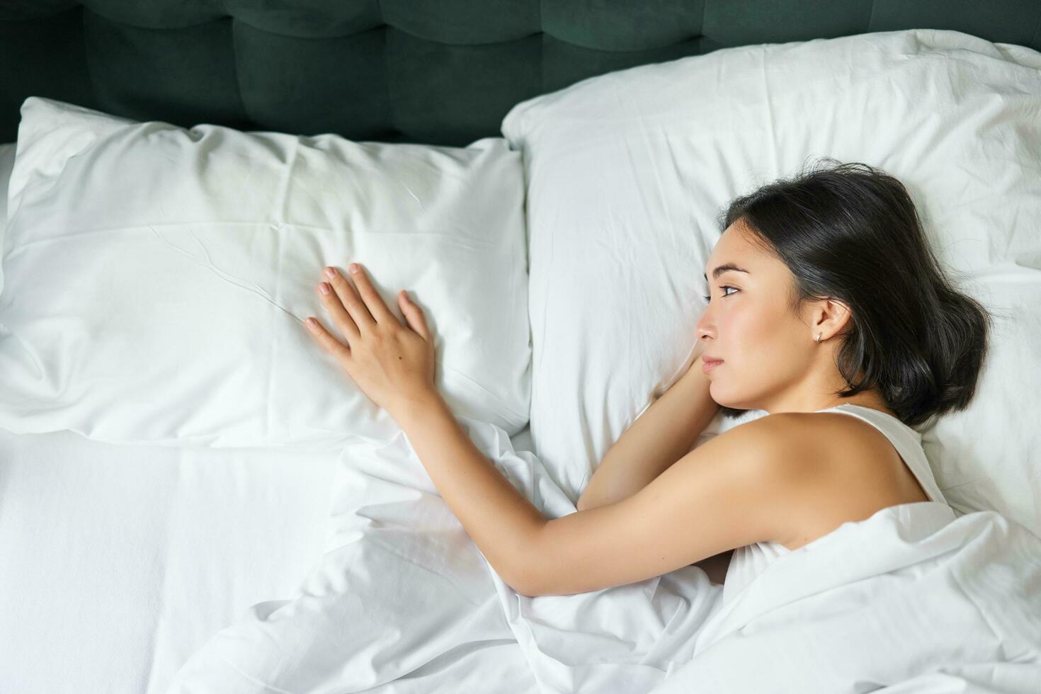 Portrait of romantic asian woman lying alone in her bed, touching empty pillow and thinking about her partner, breakup relatioship photo