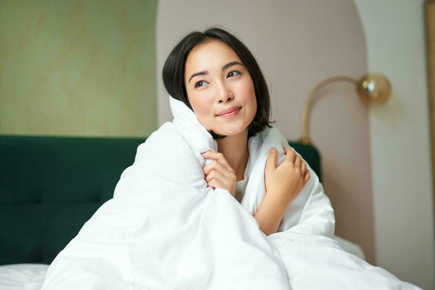 Beautiful romantic woman covers herself with warm duvet, enjoys sleeping and staying in bed, waking up happy in morning, sitting in bedroom photo