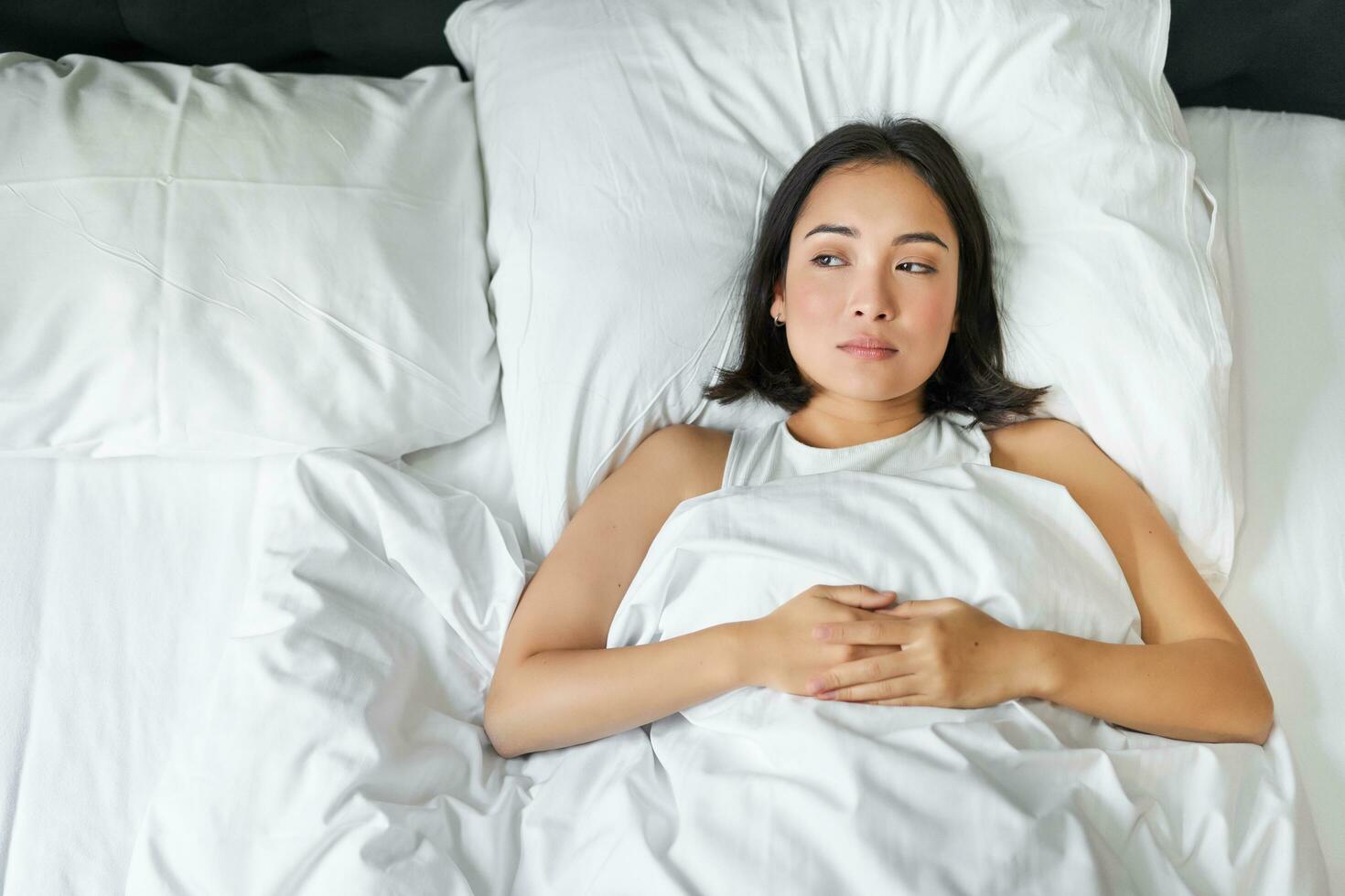 retrato de asiático niña acostado solo en cama con blanco hojas lecho, mirando considerado. mujer en su dormitorio pensando de algo y mirando a ventana foto