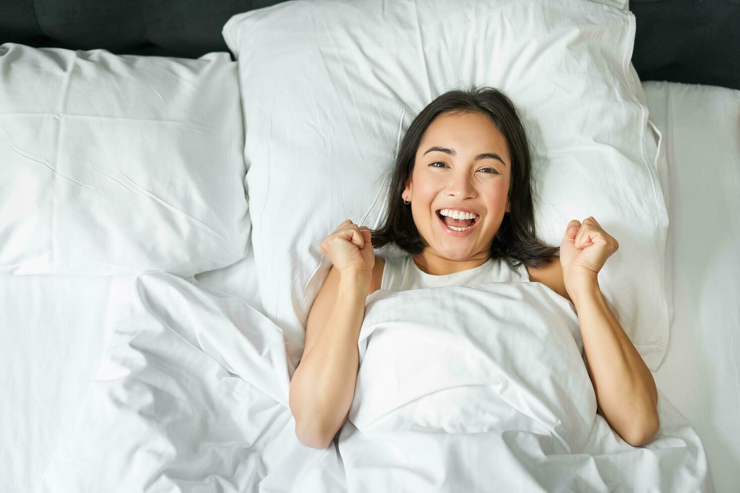 parte superior ver de entusiasta asiático niña aplausos, acostado en su cama y gritos con alegría y excitación, triunfando y sonriente, disfrutar quedarse en su dormitorio foto