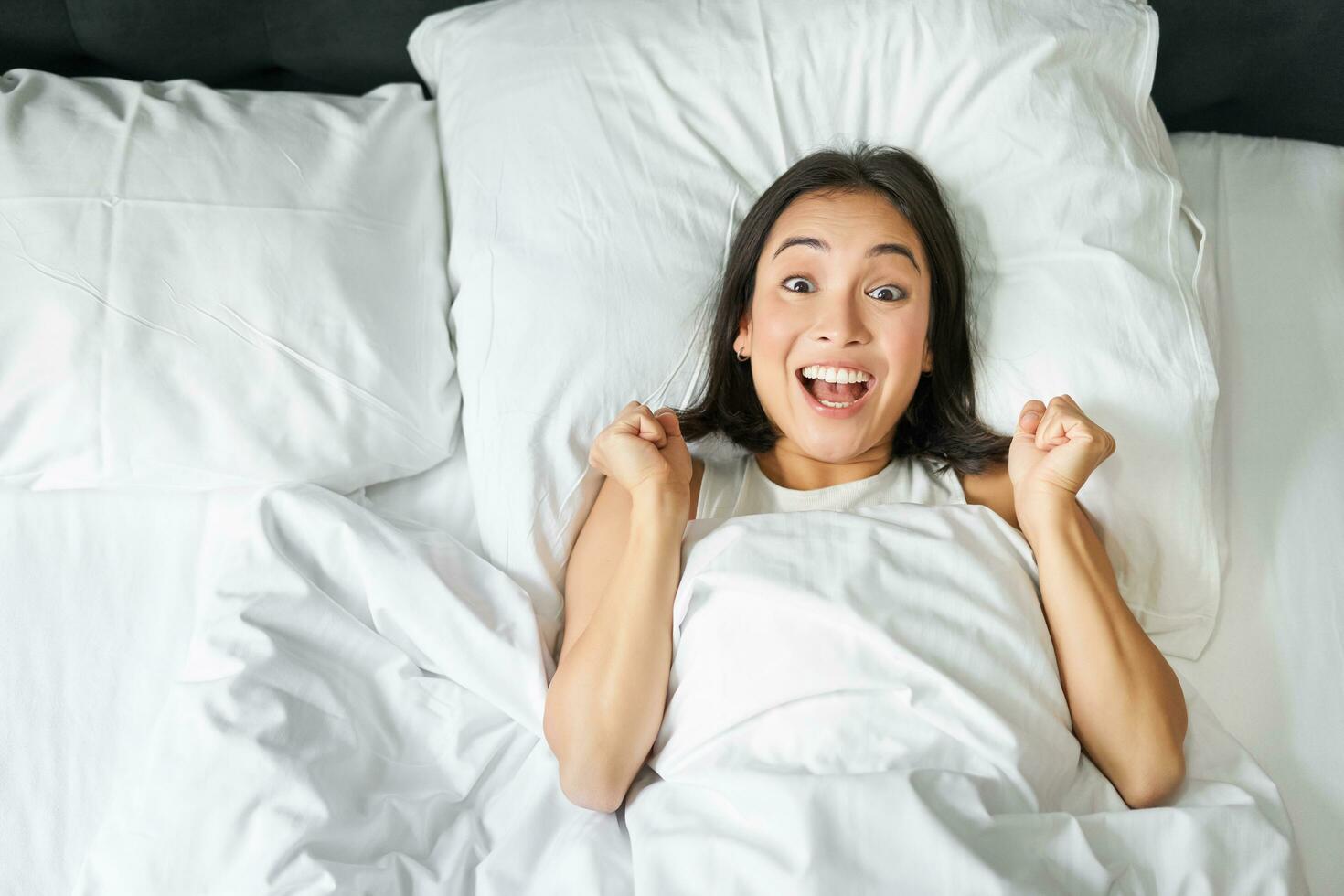 Top view of enthusiastic asian girl cheering, lying in her bed and shouting with joy and excitement, triumphing and smiling, enjoy staying in her bedroom photo