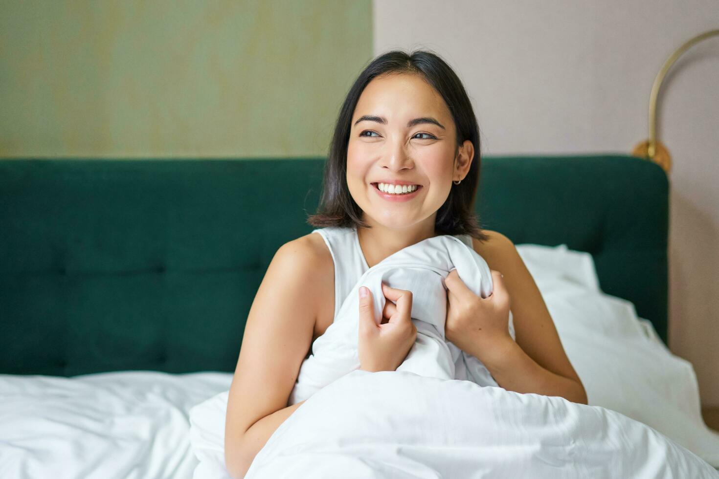 Close up of happy beautiful asian woman, waking up in bed and enjoys morning, looking outside window with sleepy smile on her face, hugging duvet photo