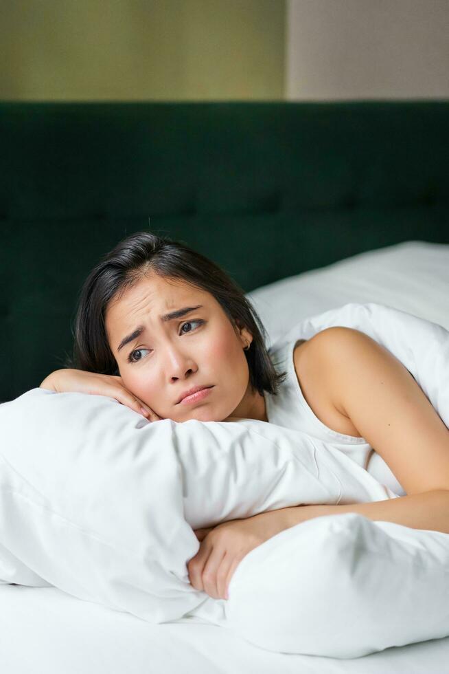 Sad and gloomy asian girl lying in her bed, grimacing and crying, feeling upset, spending time in her bedroom, hugging pillow photo