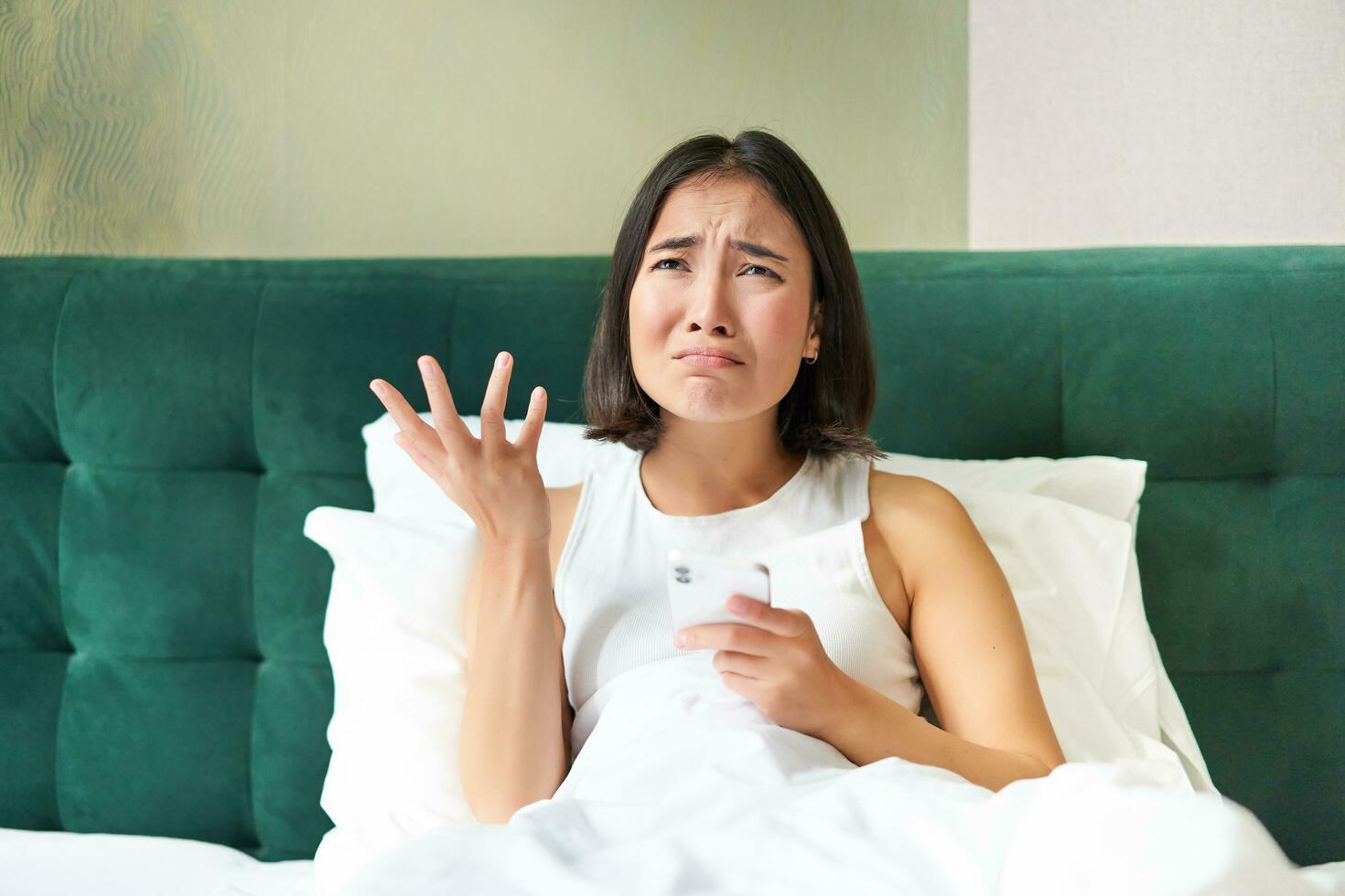 Disappointed asian girl, crying and whining, feeling upset, lying in bed on pillow, holding mobile phone, read upsetting bad news, waking up in awful mood photo
