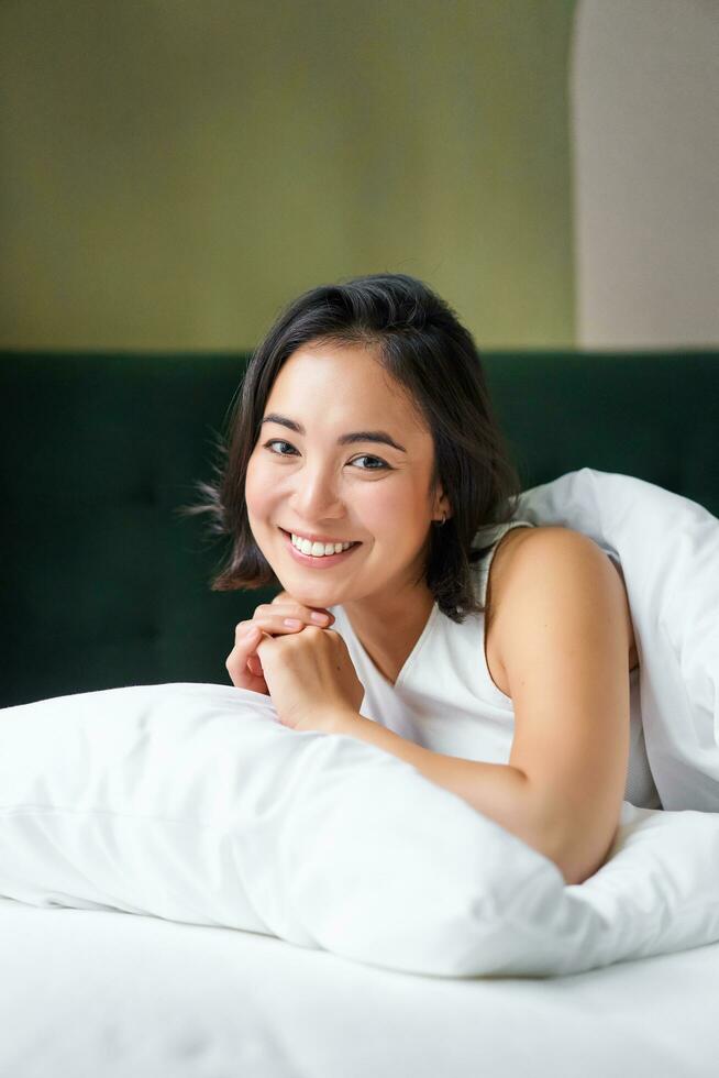 Vertical shot of smiling korean woman lying on pillow in her bed, spending morning in bedroom, waking up from sleep, looking happy photo