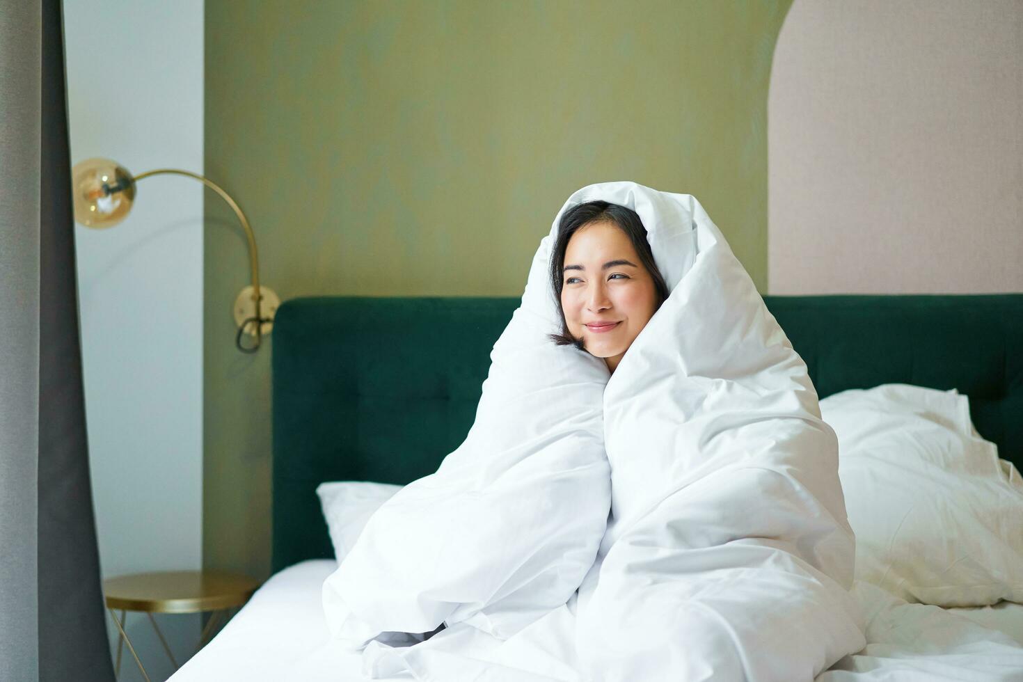 Cozy mornings. Happy asian girl feels warm in her bed, covers herself with cozy duvet in her bedroom, a comfortable stay in hotel room photo