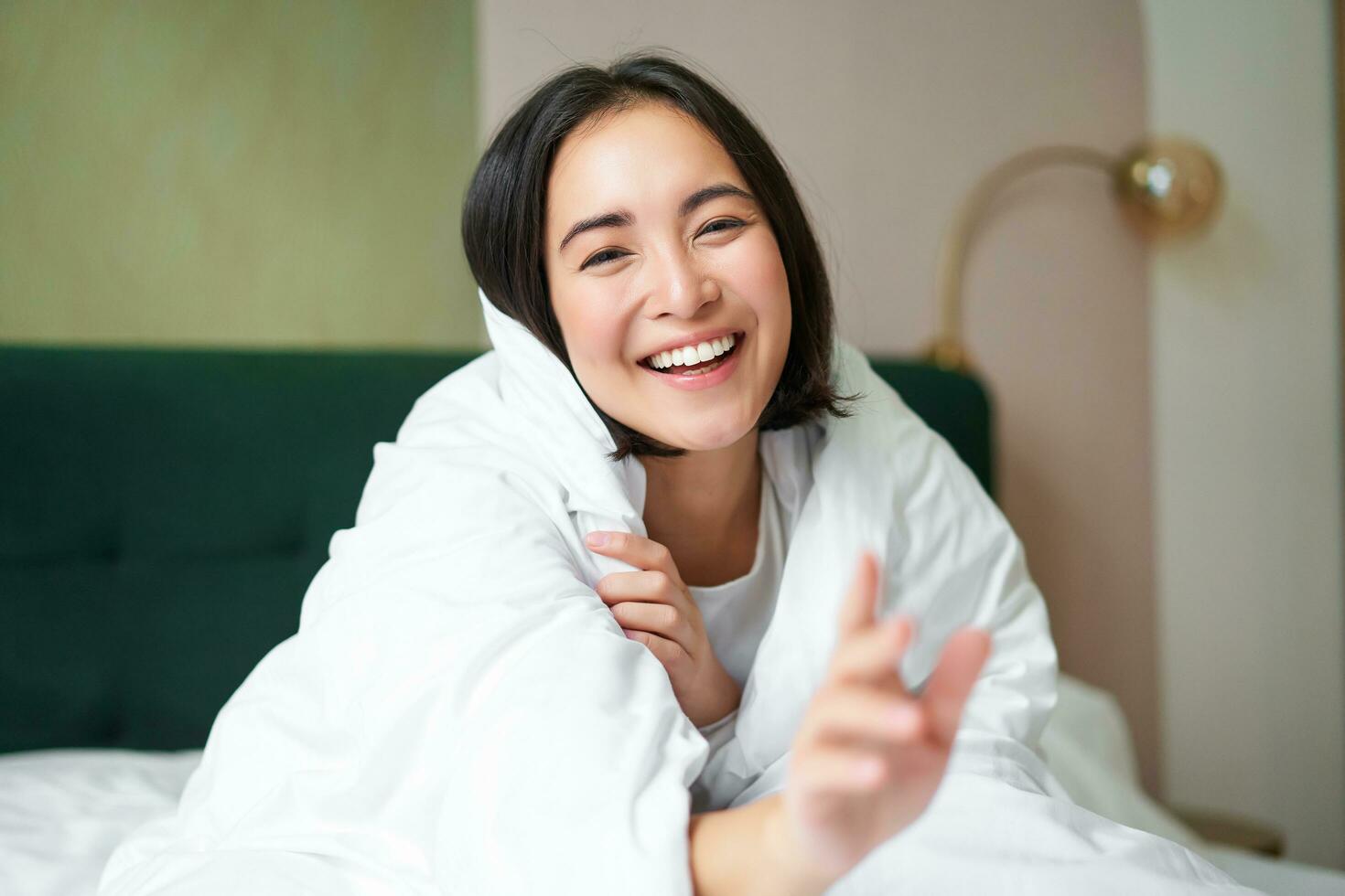 Happy beautiful asian woman covered with blanket, sits on bed in warm duvet, tries to reach camera with a hand, smiling playfully. Hotel bedroom, comfort and apartments concept photo