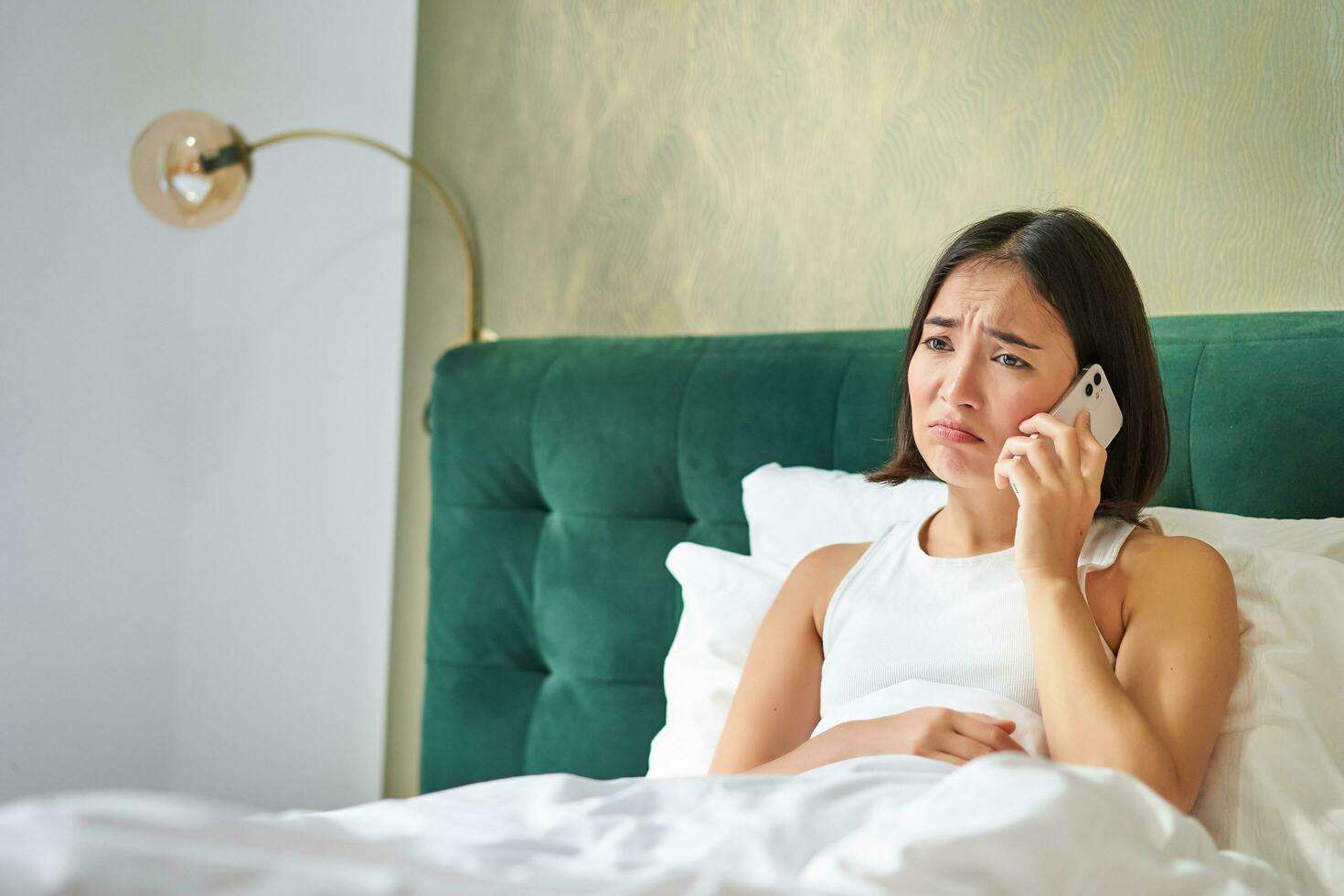 retrato de preocupado asiático mujer participación móvil teléfono, recibir malo teléfono llamar, mirando preocupado y decepcionado, teniendo difícil teléfono conversacion mientras acostado en cama foto