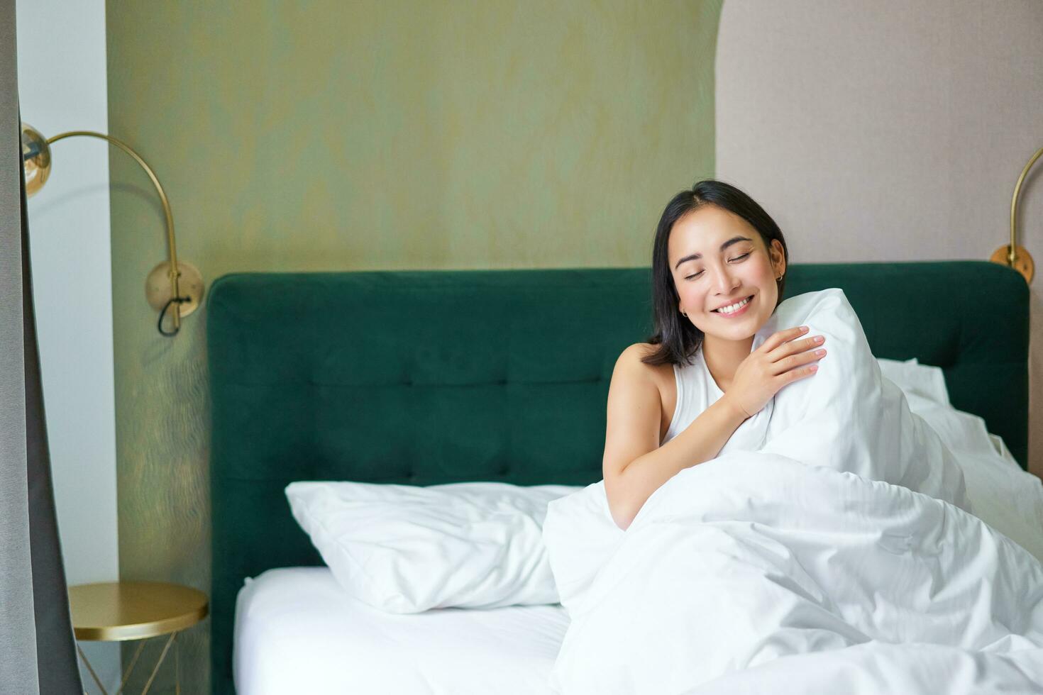 Happy korean woman wakes up in her hotel room, lying in bed under cozy warm blankets, white sheets, enjoying weekend morning photo