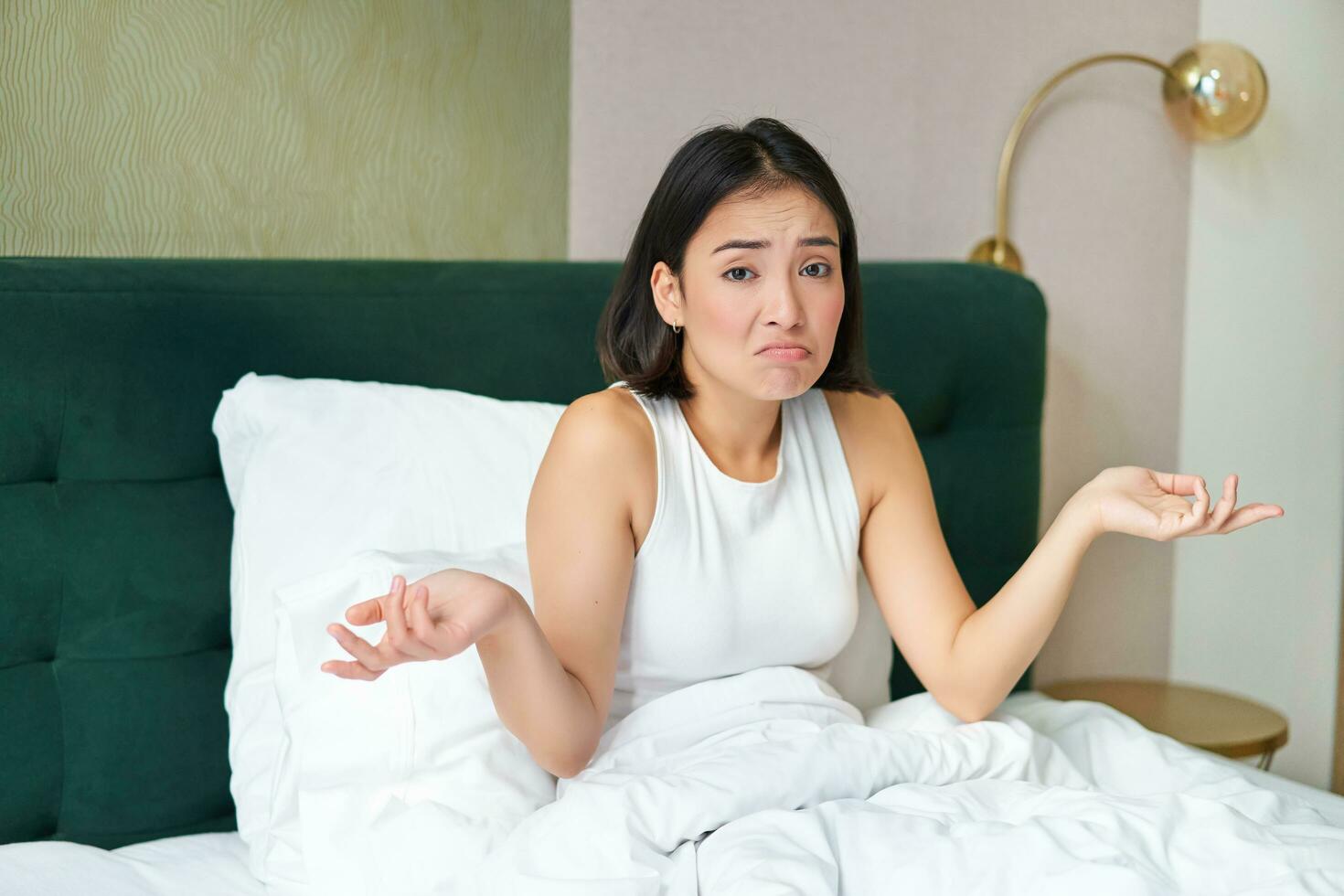 Confused asian girl lying in bed and shrugging shoulders, looking puzzled, dont know, spending time in her bedroom photo