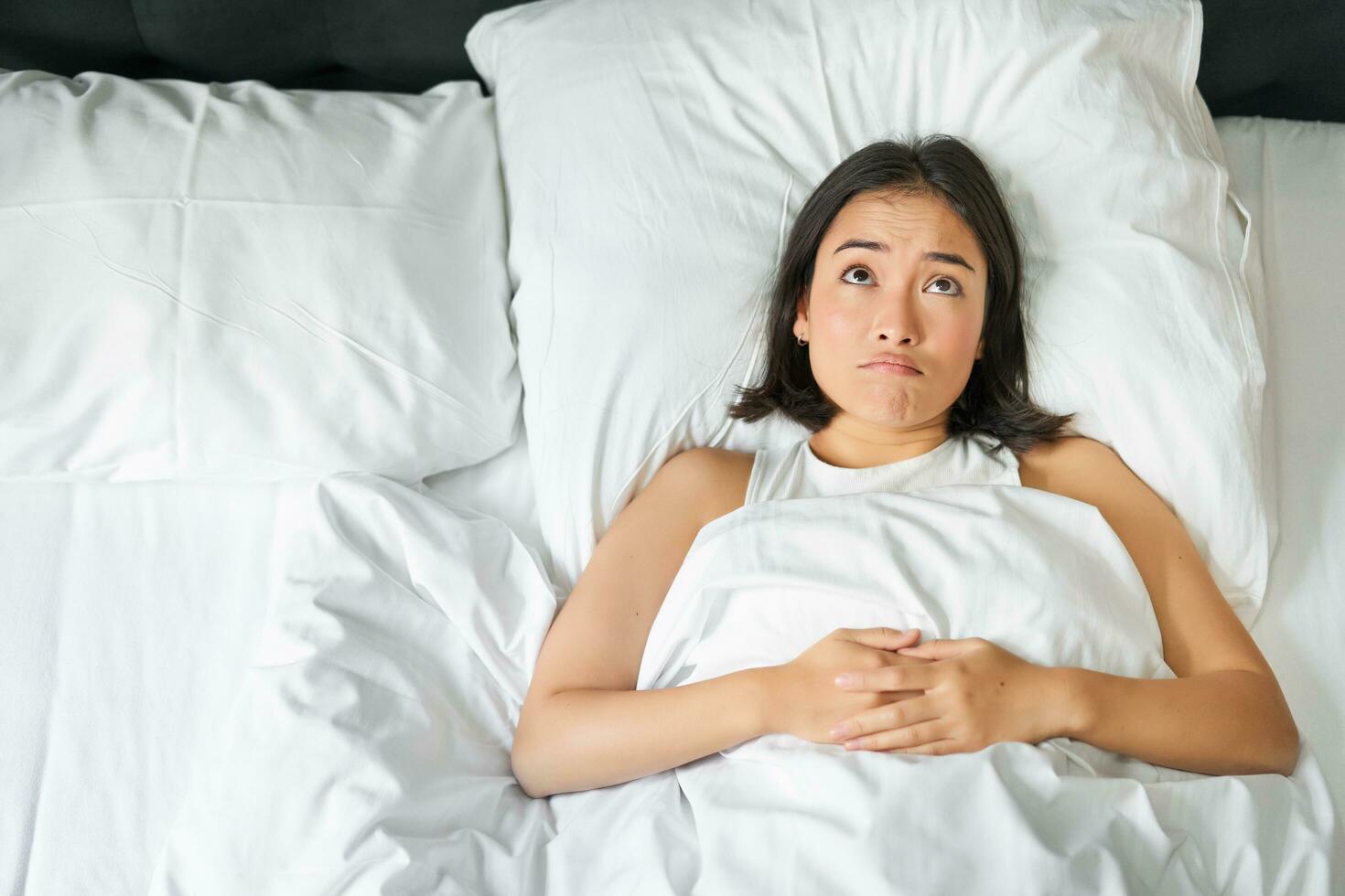 Portrait of asian girl lying in her bed and shrugs, looks puzzled, overthinking before going to sleep, looking complicated at camera photo