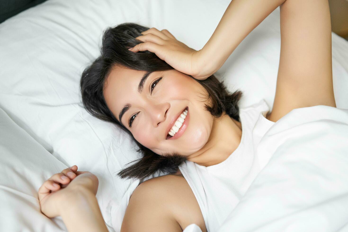 cerca arriba de contento asiático niña acostado en cama en su almohada, sonriente y laminación terminado como sensación energético en mañana, despertar arriba temprano foto