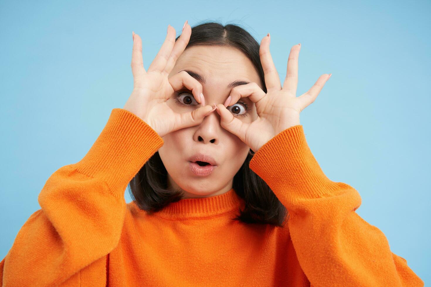 Close up of funny korean girl, shows glasses and makes silly face, stands over blue background photo