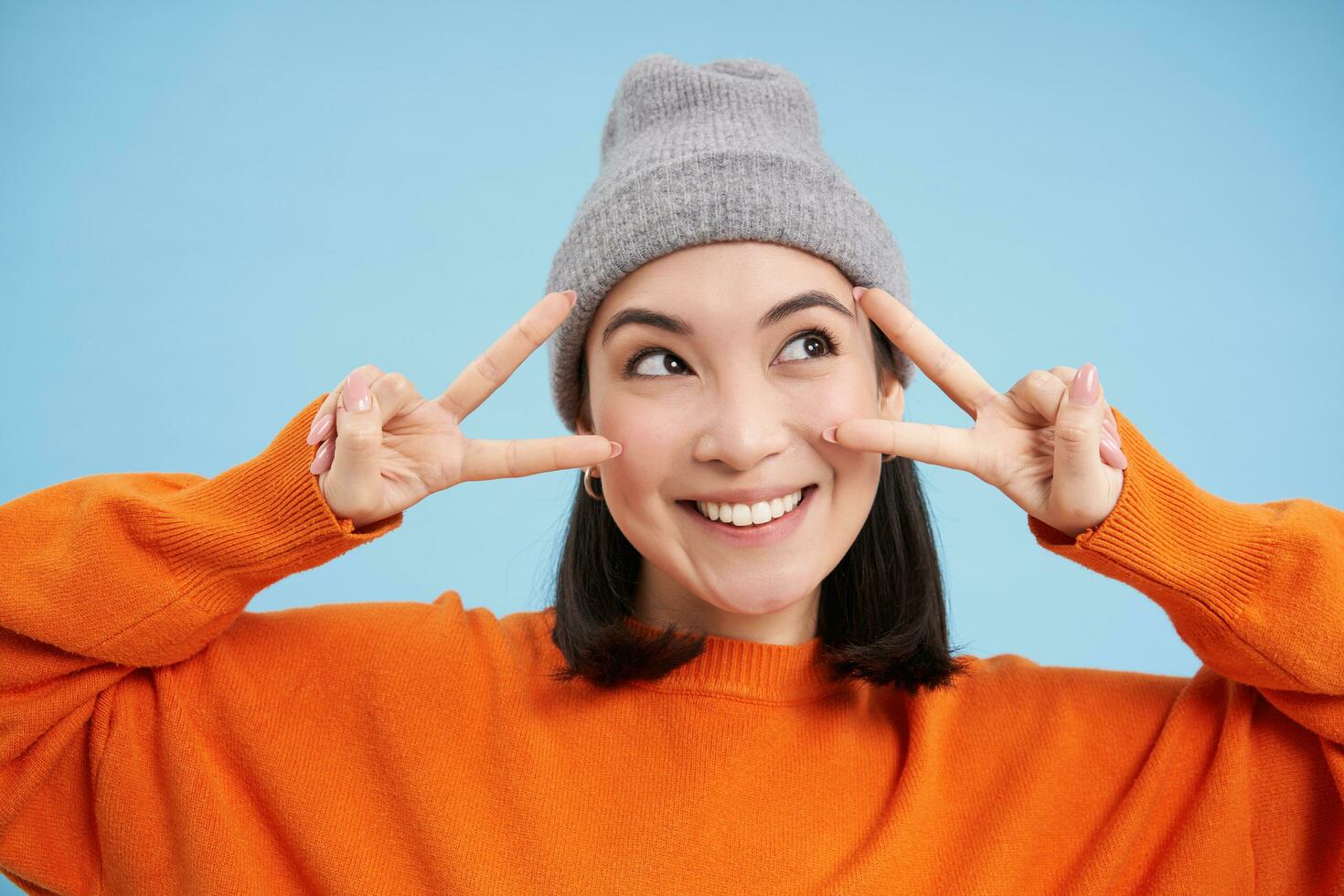 Close up portrait of cute korean girl in beanie, shows peace, vsign gesture, positive vibe, smiling and laughing, posing over blue background photo